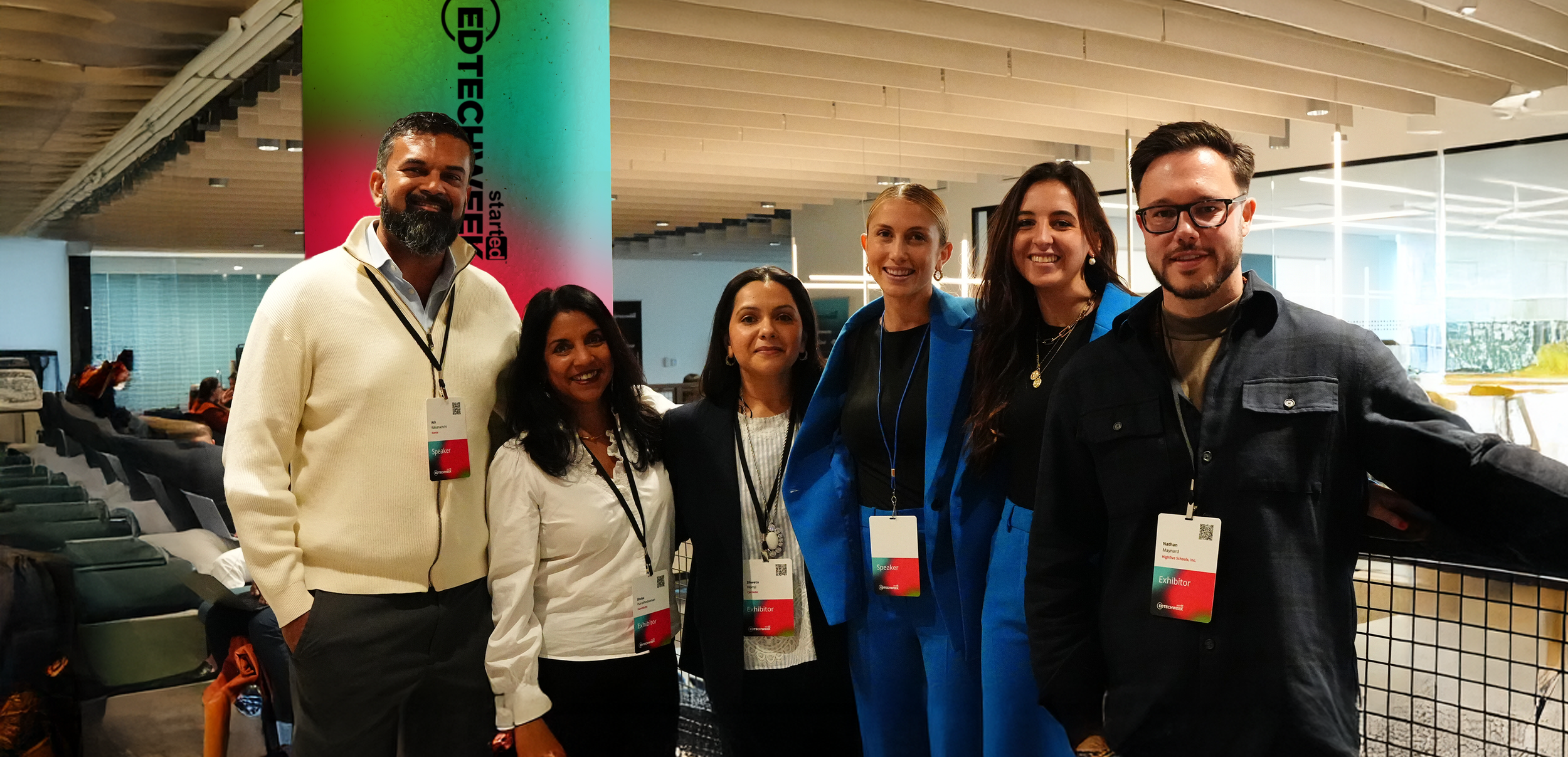 Group of six diverse professionals at conference, wearing badges, standing indoors with seating and a colorful vertical banner in the background.