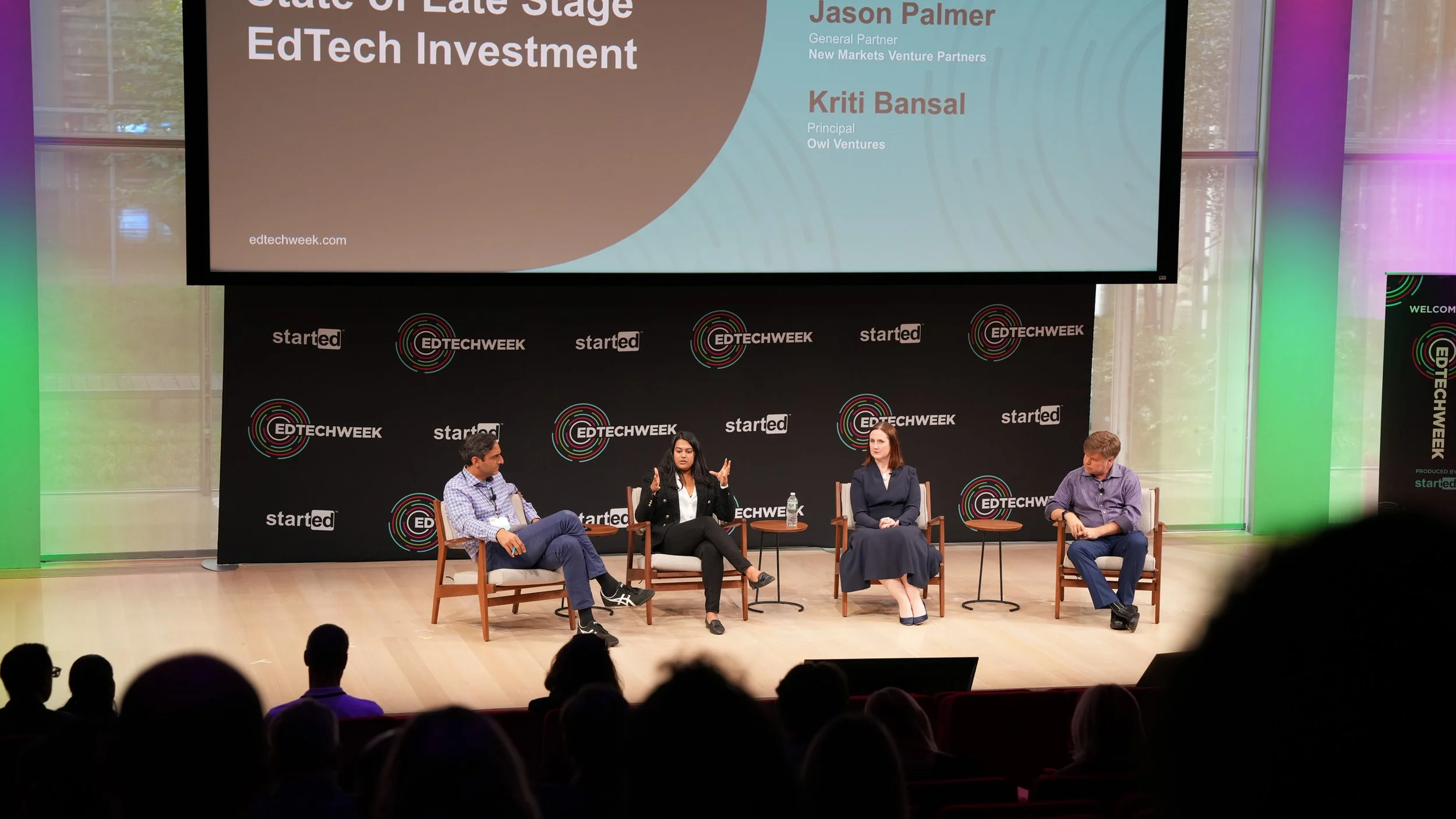 Panel discussion at EdTech Week, featuring four speakers seated on stage with a large screen behind them displaying titled panel discussion, and a backdrop with 'EDTECH WEEK' logos.