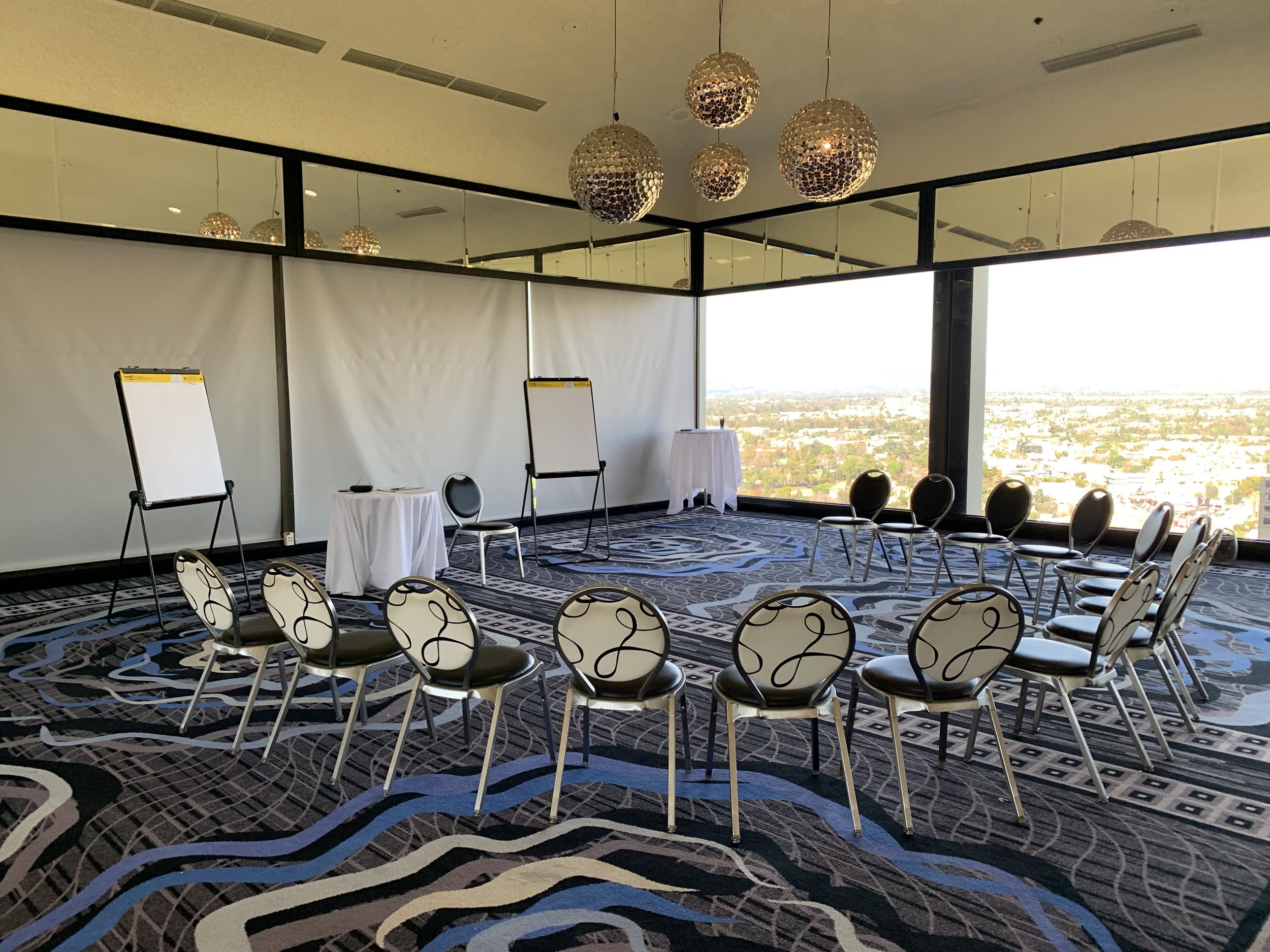 Set up for a meeting or seminar in a high-rise conference room with large windows overlooking a cityscape, featuring chairs arranged in a semi-circle, whiteboards, and a table with a tablecloth.