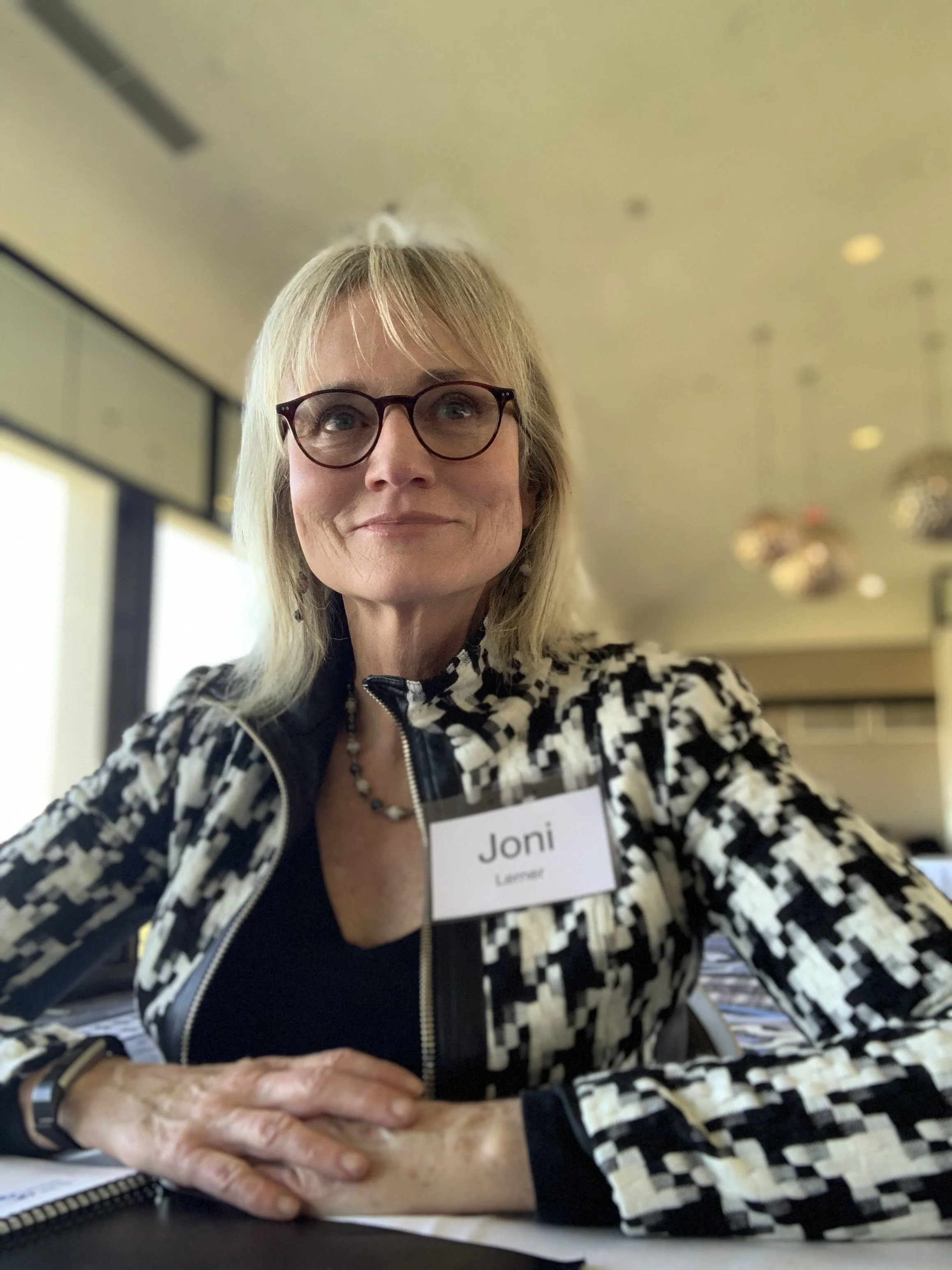 A woman with blonde hair, glasses, and a black and white patterned jacket sitting at a table, wearing a name tag that reads 'Joni Lerner', inside a modern conference room with ceiling lights.