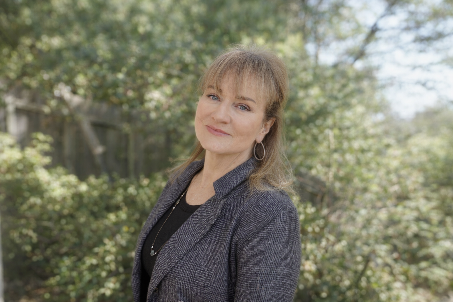A woman with shoulder-length light brown hair and blue eyes, wearing a black top and a gray checkered blazer, standing outdoors in a natural setting with green trees and sunlight.
