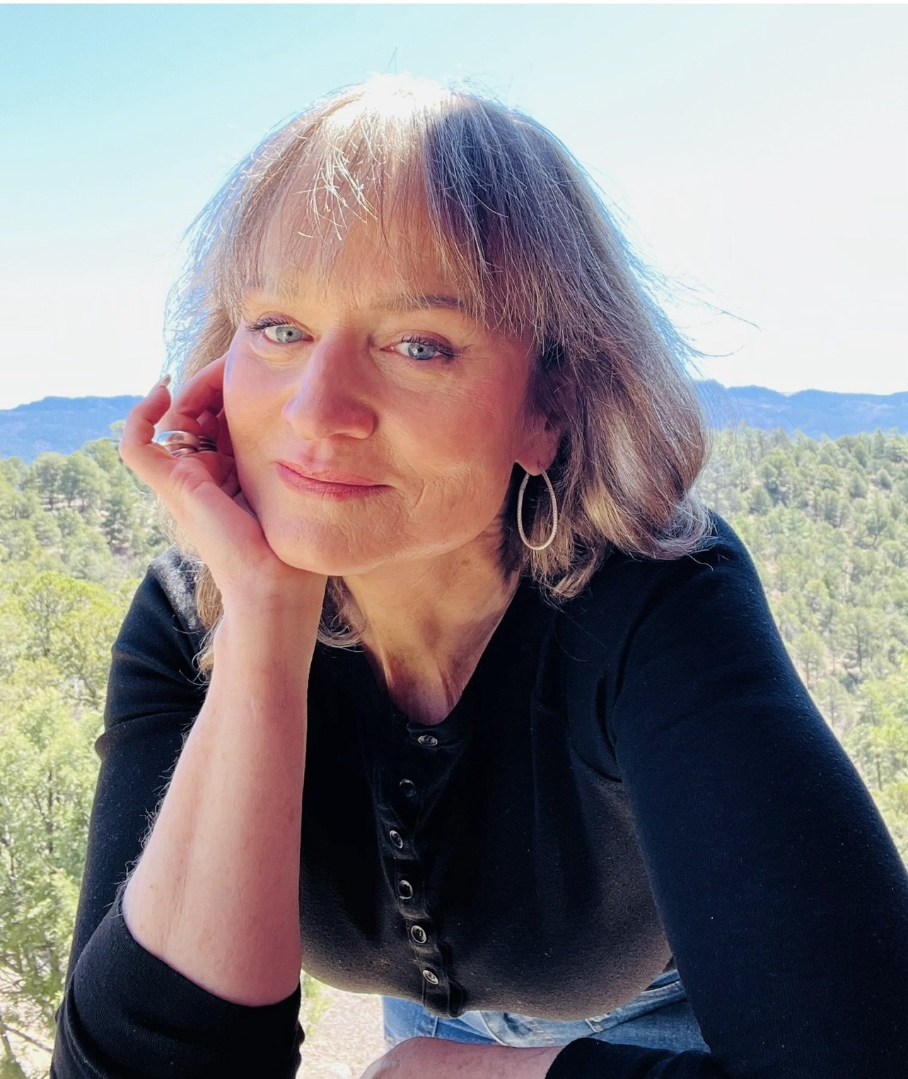 A woman with short light brown hair and blue eyes wearing a black shirt and silver hoop earrings, outdoors with a landscape of trees and mountains in the background.