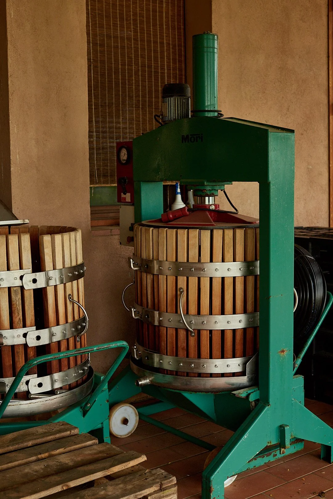 A traditional wooden wine or grape press machine with a green metal frame and two barrels, placed indoors against a beige textured wall.