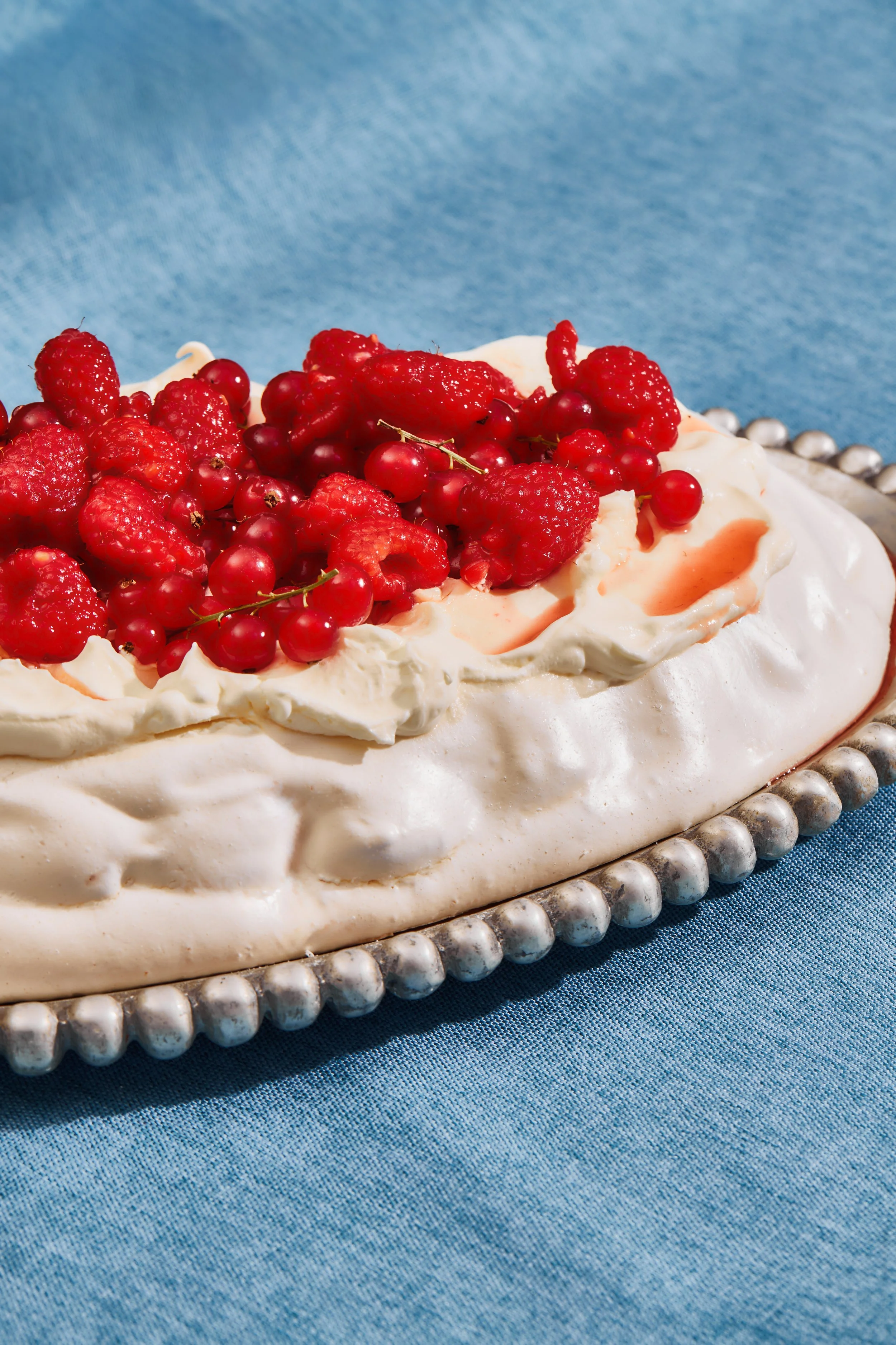 A pavlova dessert topped with whipped cream and mixed berries, including raspberries and red currants, on a silver platter with a blue cloth background.