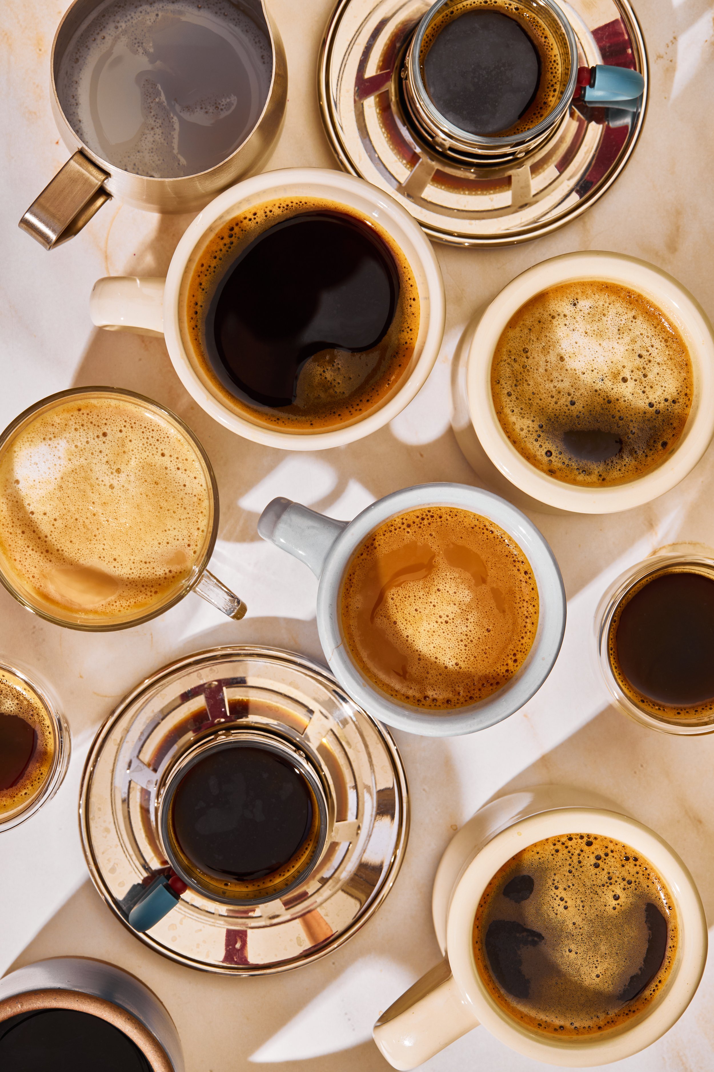 Multiple cups and mugs filled with black coffee on a table, some with foam, along with a few glasses containing dark beverages.