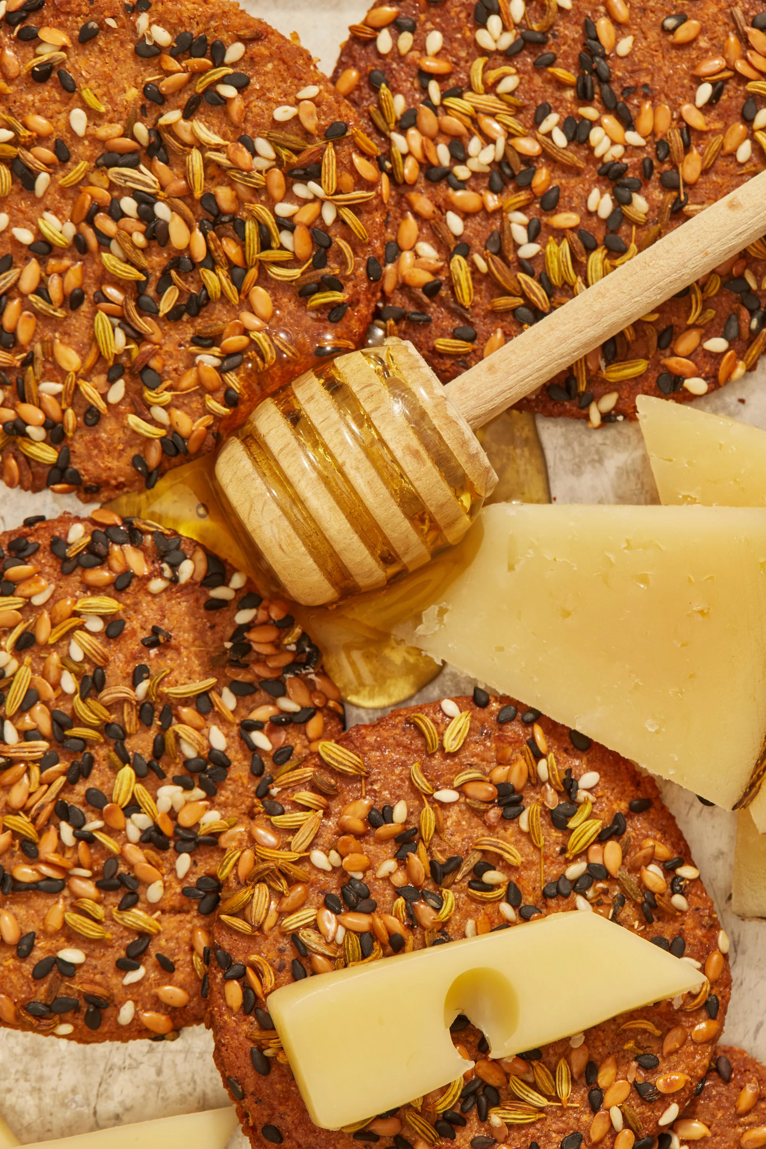 Close-up of honey glazed cookies topped with mixed seeds, with slices of cheese and a honey dipper.