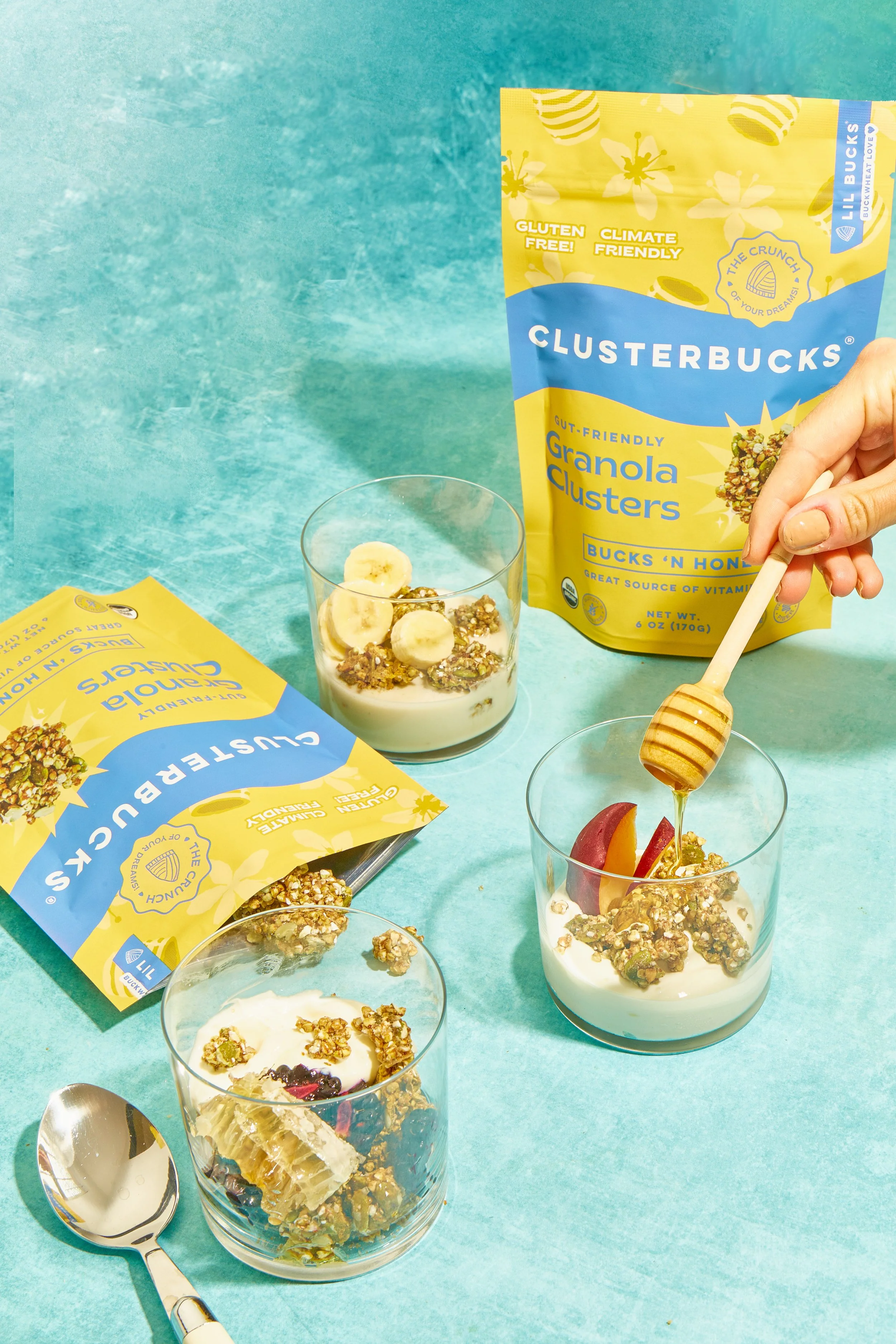 Close-up of a breakfast scene with bowls of yogurt topped with granola, sliced bananas, and peach slices, along with a bag of ClusterBucks granola clusters and a spoon with honey.