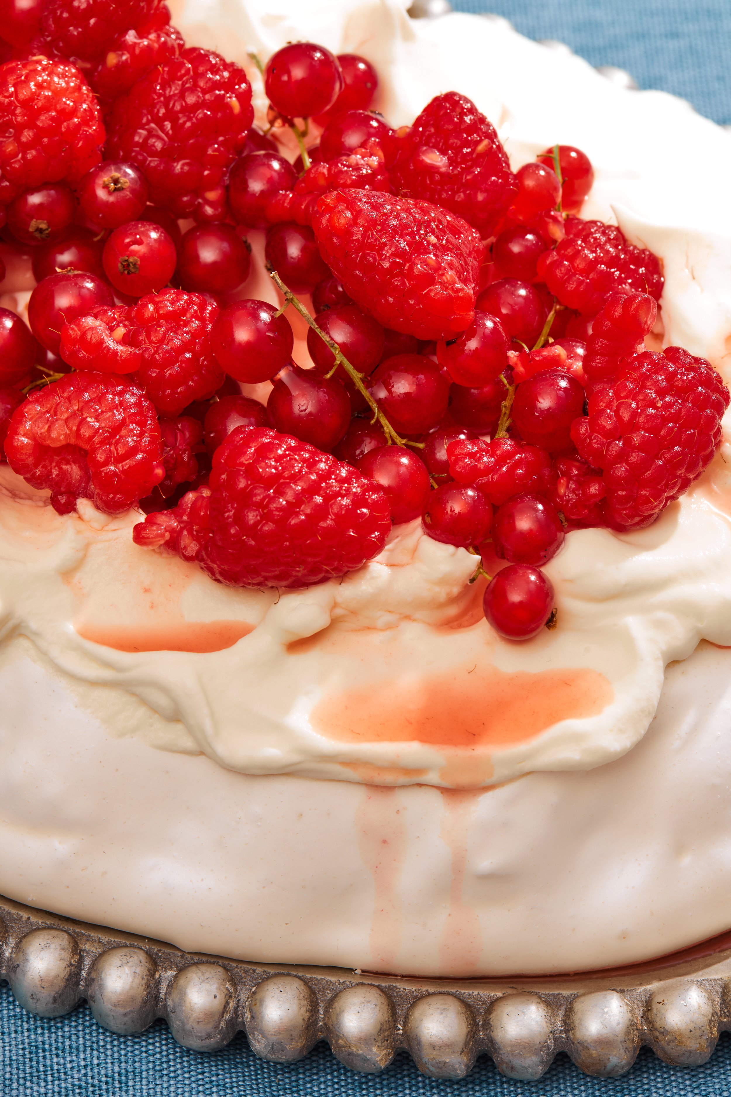 A close-up of a dessert topped with fresh raspberries and redcurrants, with a dollop of whipped cream and a light drizzle of syrup, on a silver cake stand with a beaded edge.