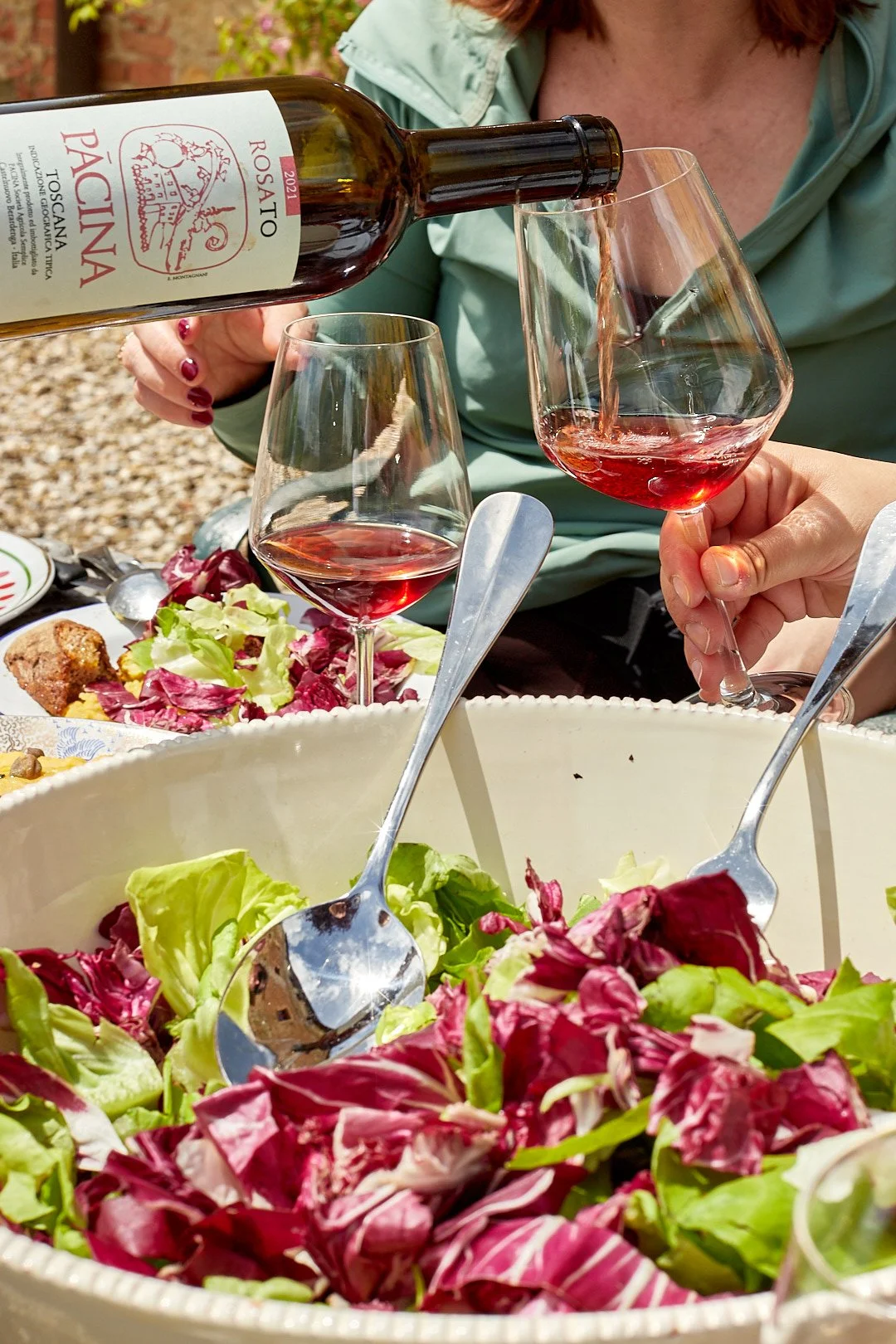 Person pouring red wine into a glass at a table with salad and other food items outdoors.
