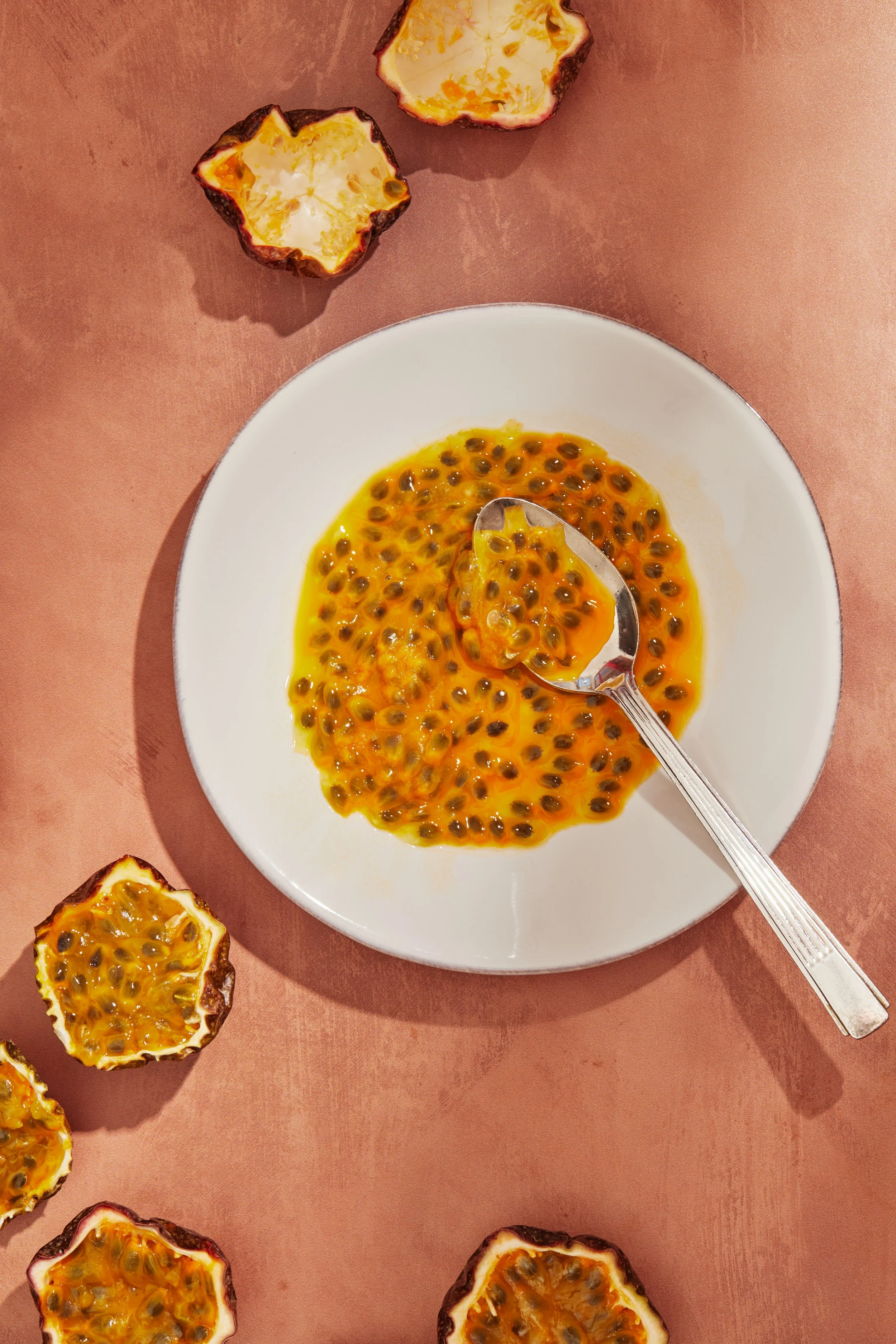 A white plate of passion fruit pulp with seeds and a spoon resting inside, with halved passion fruits around the plate on a pink surface.