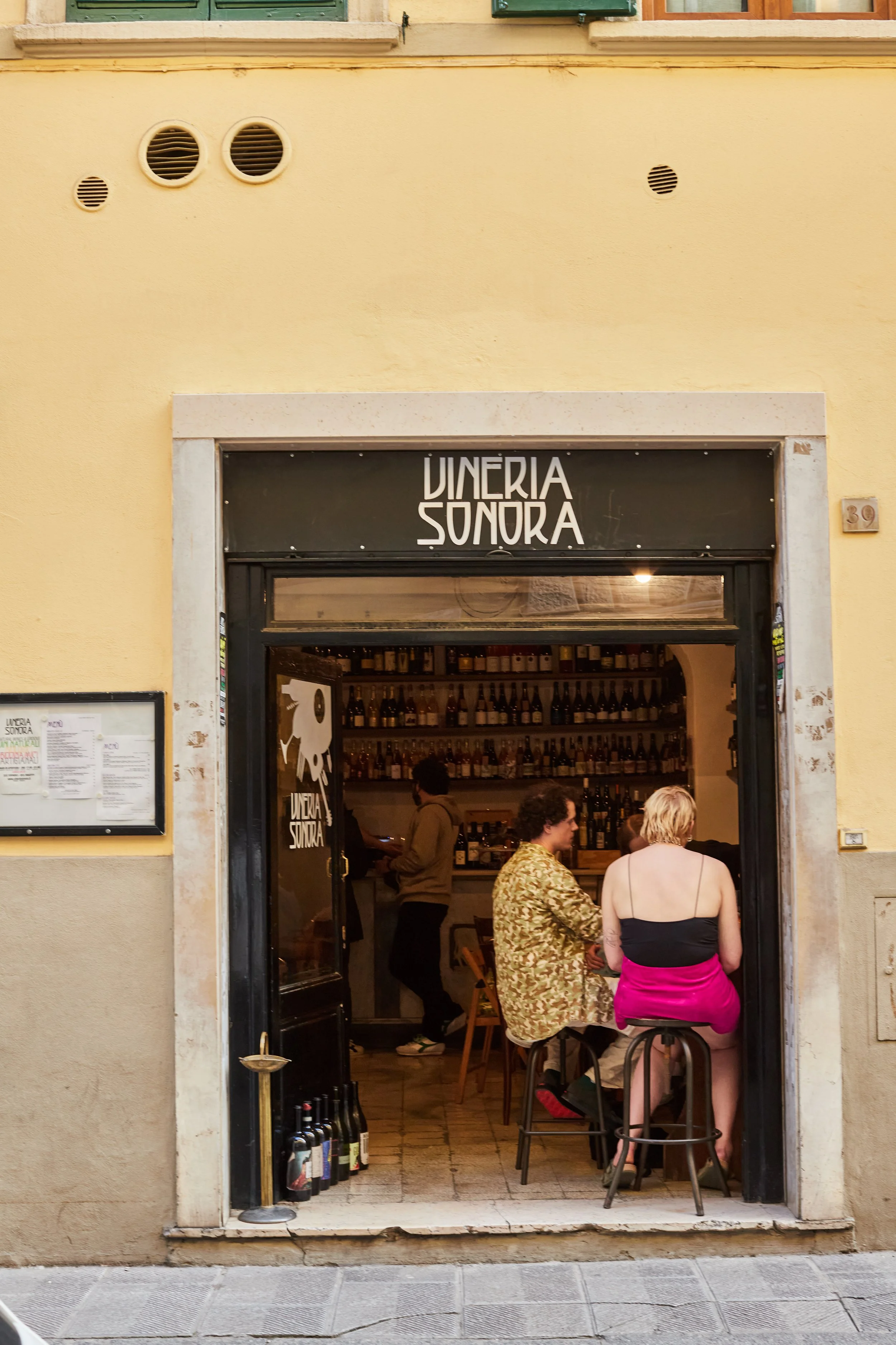 Entrance of a Florence wine bar named 'Vineria Sonora' with customers inside and outside, wine bottles visible near the doorway, and people seated at a table through the open door.