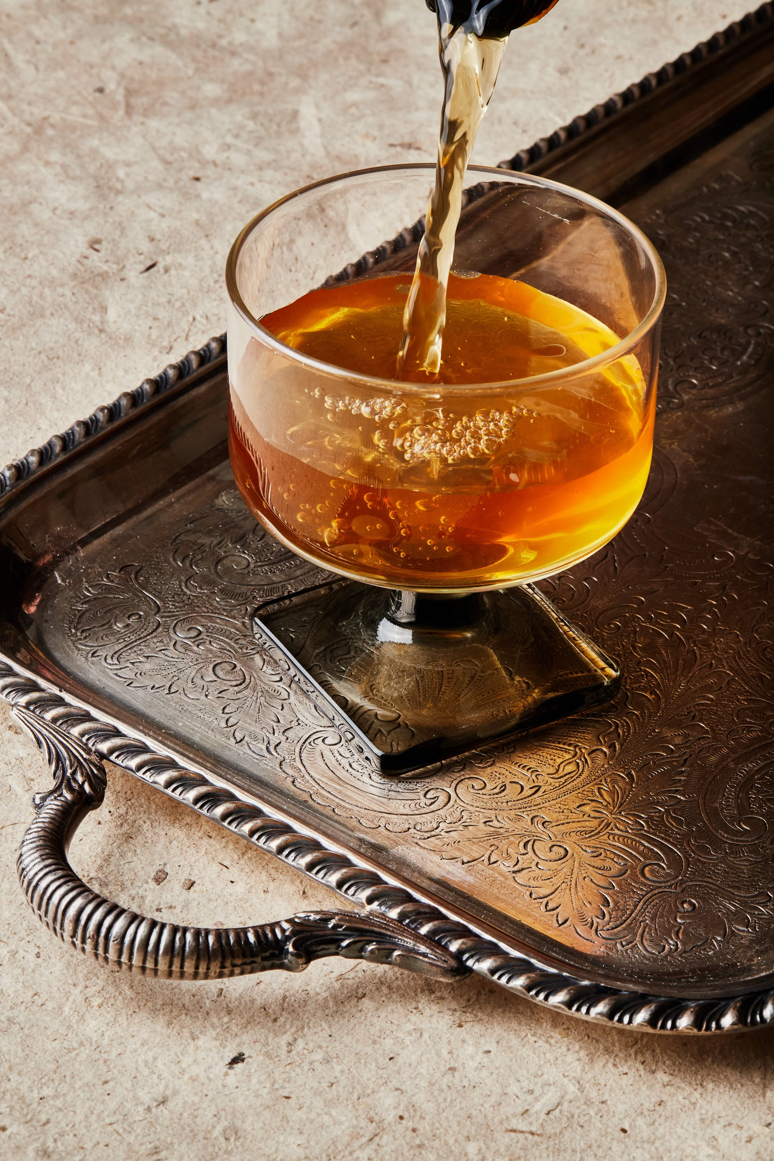 A glass of wine is being poured on a decorative metal tray with intricate patterns.