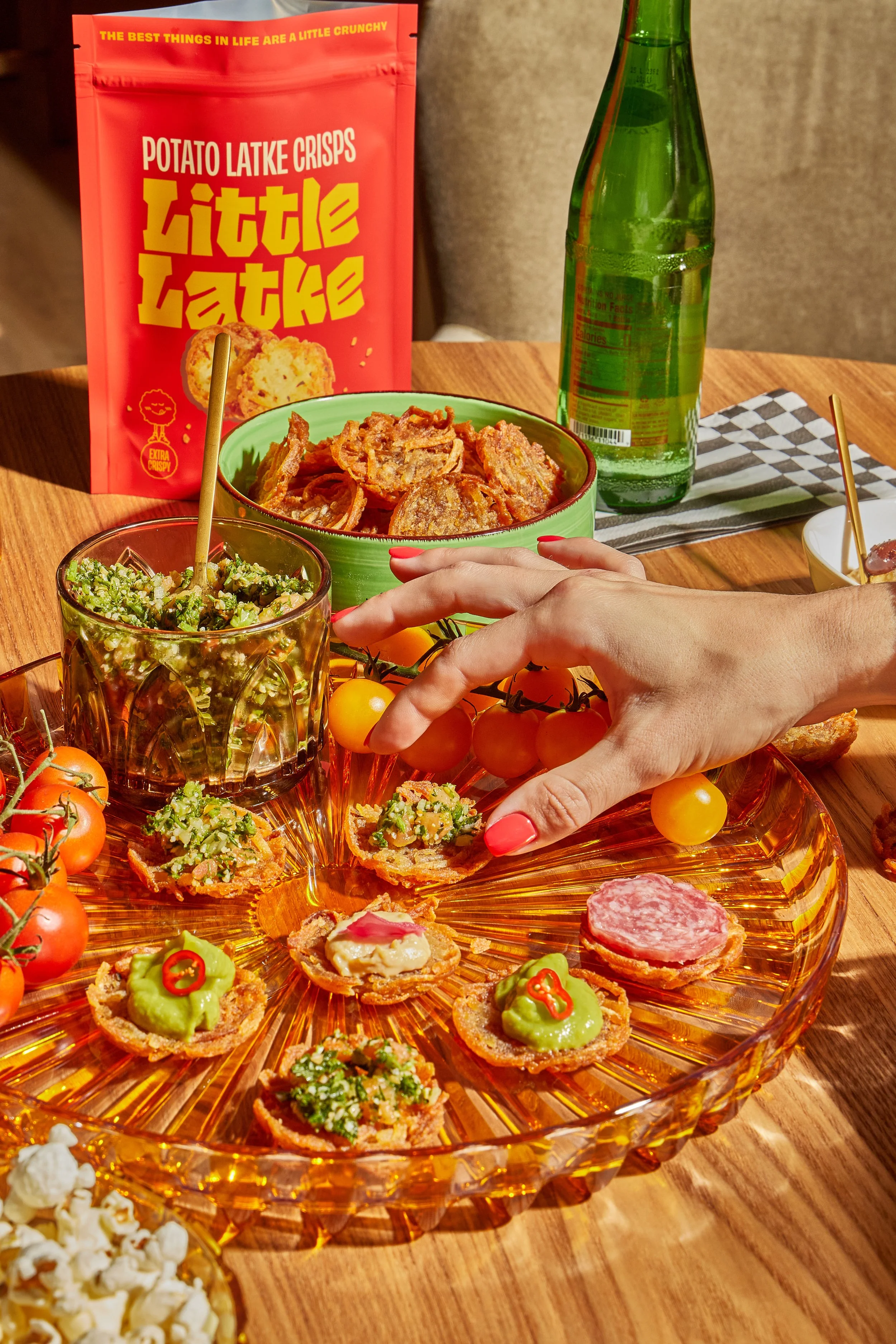 A woman reaching to pick an appetizer from a transparent, decorative orange glass serving platter filled with various appetizers including guacamole, salami, and other toppings. The table also has cherry tomatoes, a glass bottle of soda, a bowl of po