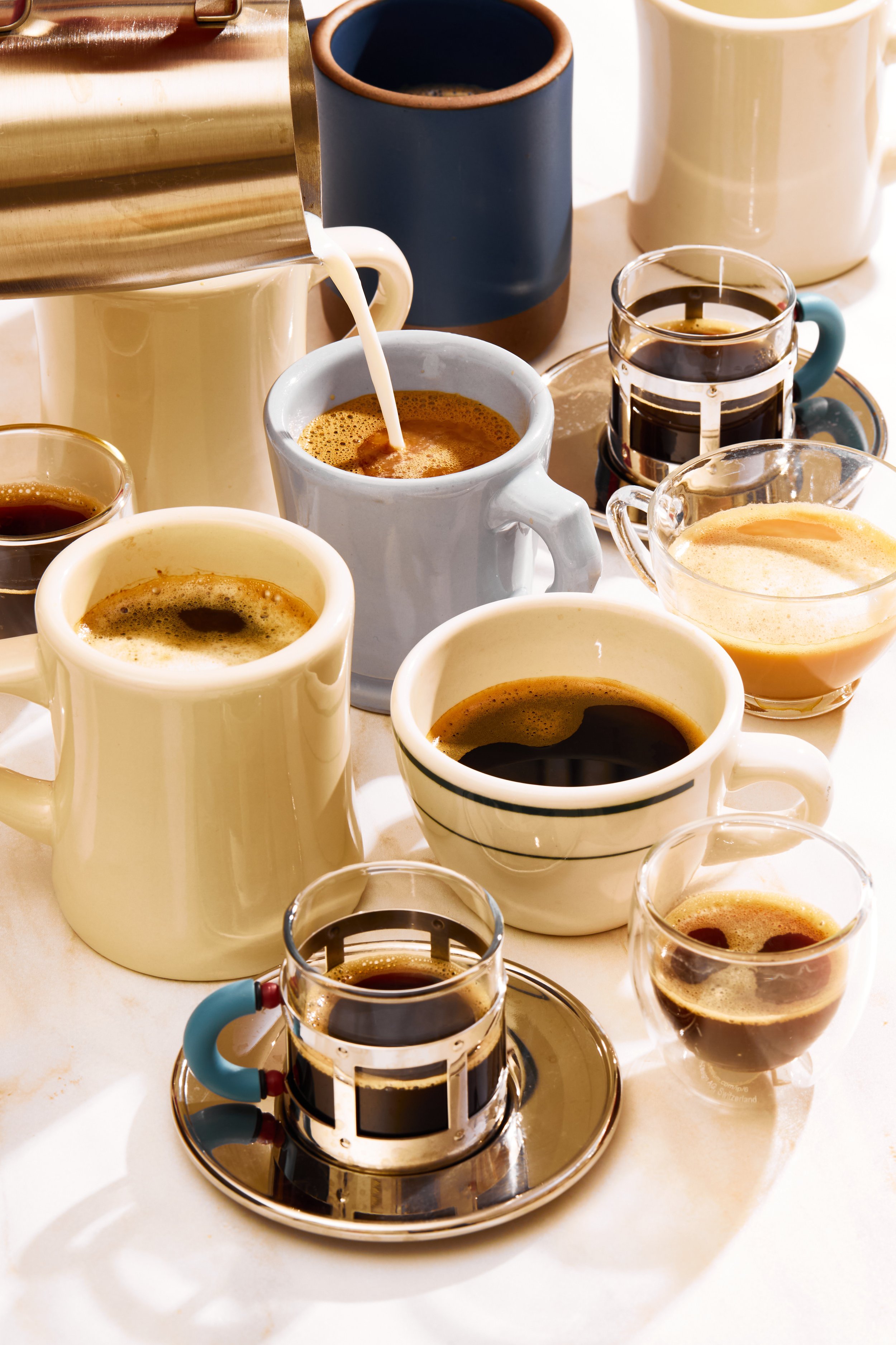 Multiple cups and glasses of black coffee and espresso on a table, with a milk frother pouring milk into one cup.