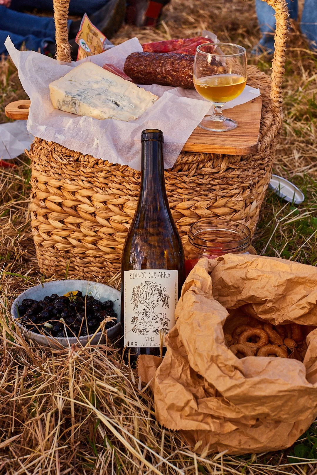 A picnic setup on the grass with a wicker basket, various cheeses, a glass of white wine, a bottle of wine labeled 'Bianco Susanna', a bowl of black olives, a jar of jam, and a crumpled paper bag containing small bread rings.