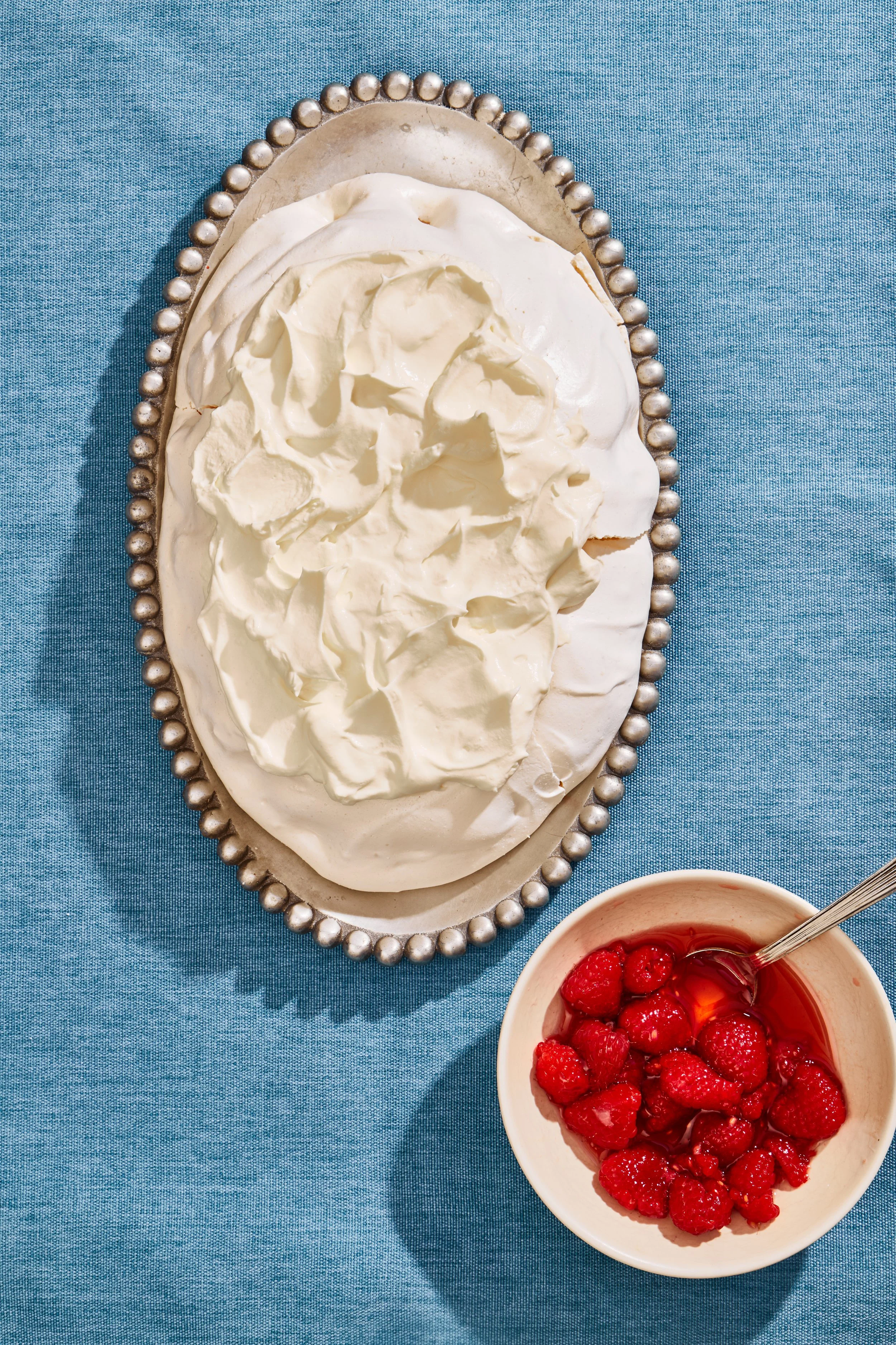 A bowl of raspberries with syrup and a spoon, placed next to an ornate silver serving dish filled with whipped cream on a blue tablecloth.
