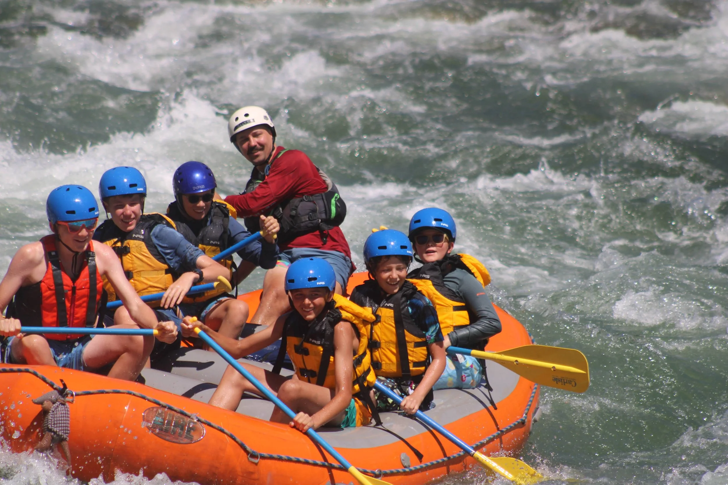 Group of people white water rafting, wearing helmets and life jackets, paddling and splashing in rushing river water.