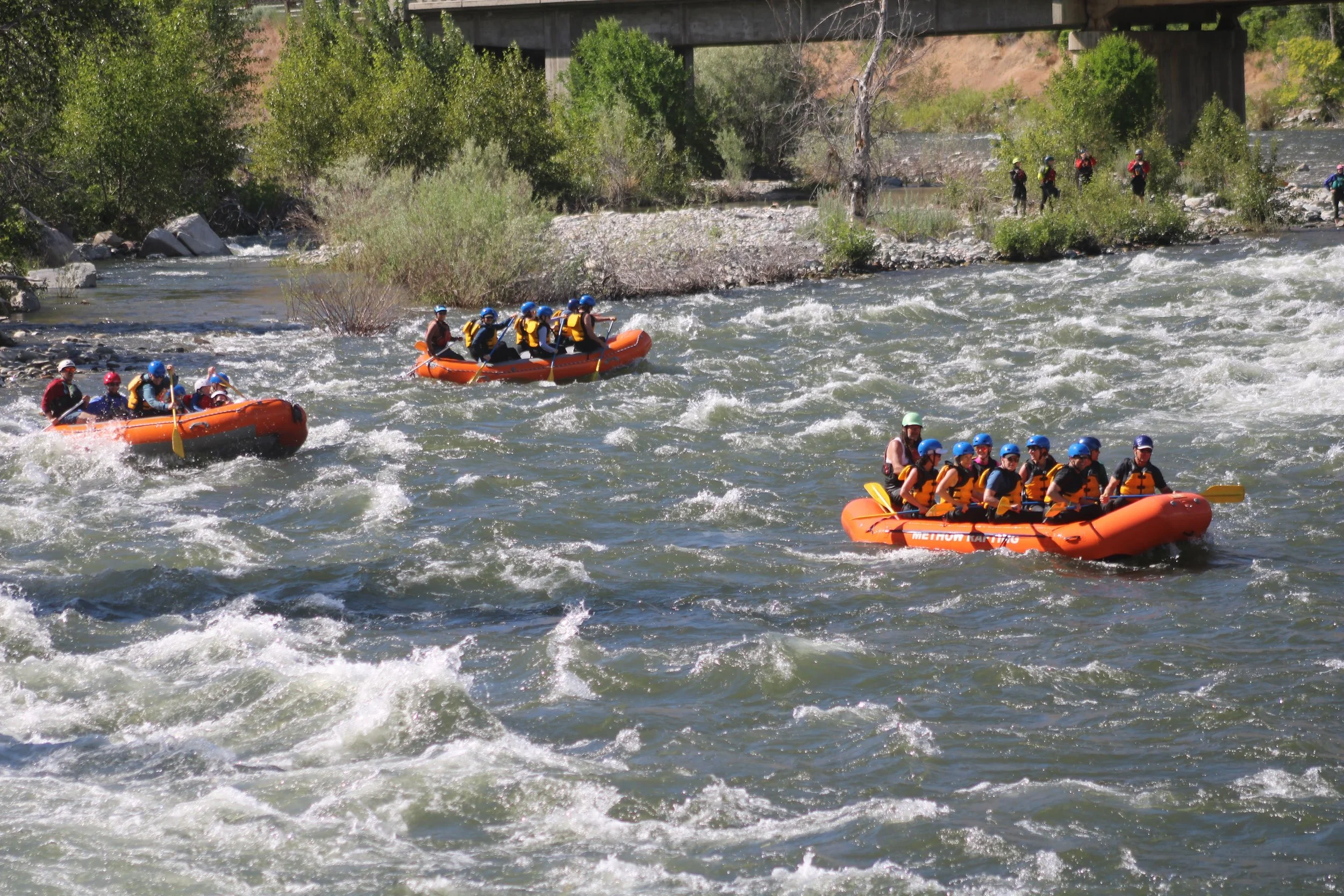 Group of people in orange inflatable raft on a river with trees and hills in the background, under a blue sky with some clouds.
