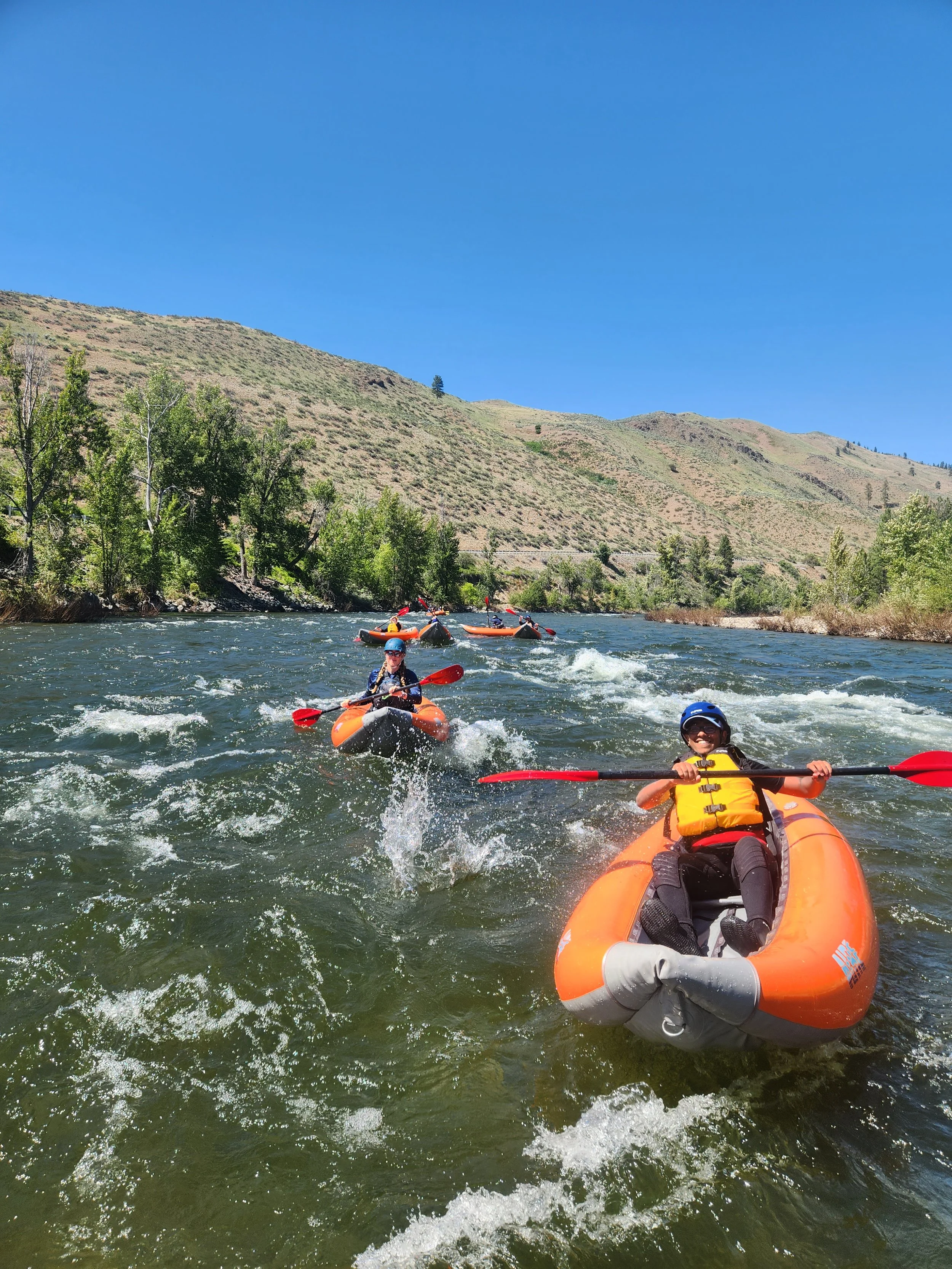 People kayaking on a river with green trees and hills under a clear blue sky.