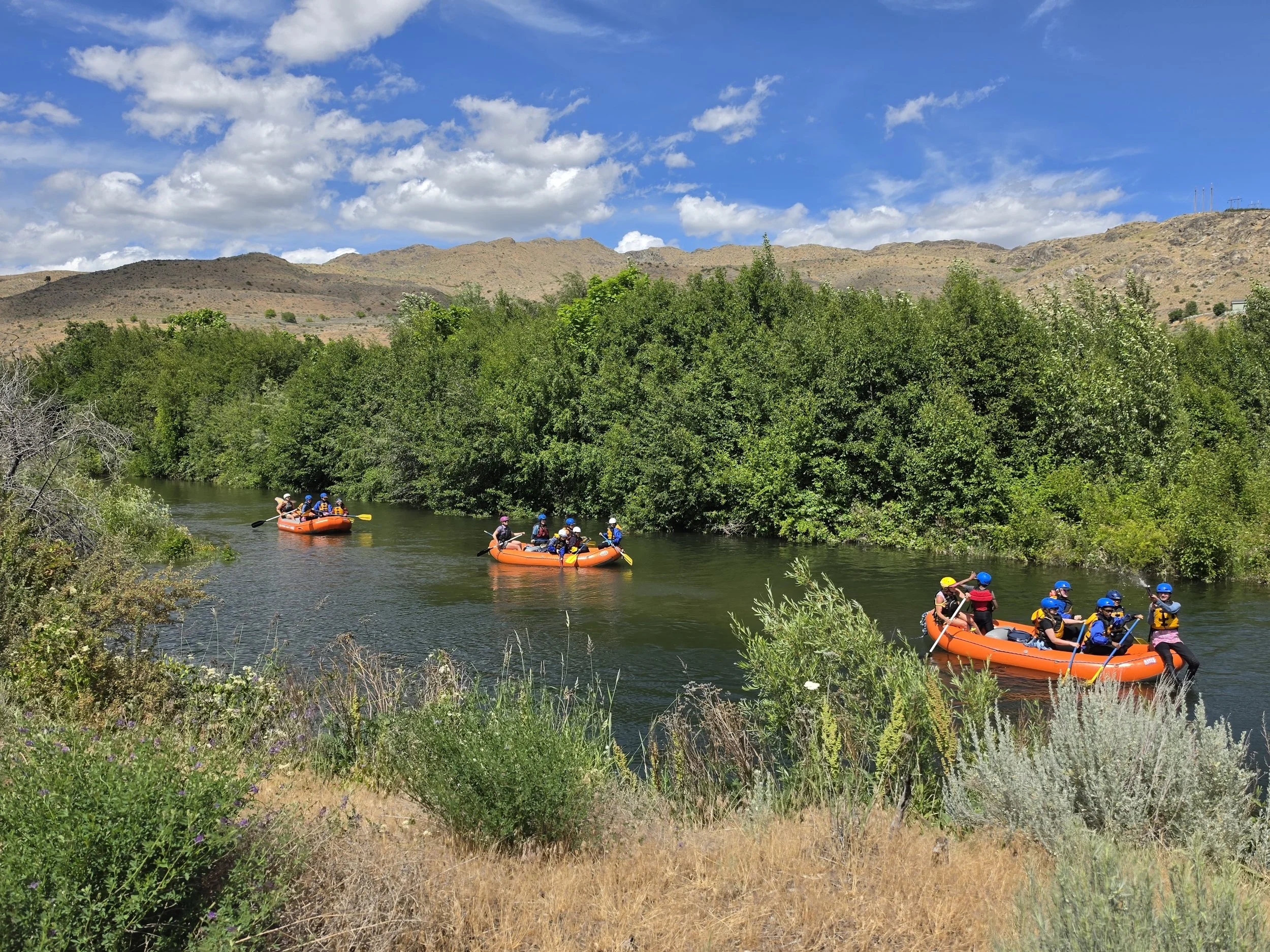 Group of people rafting on a river surrounded by trees and mountains under a clear blue sky.