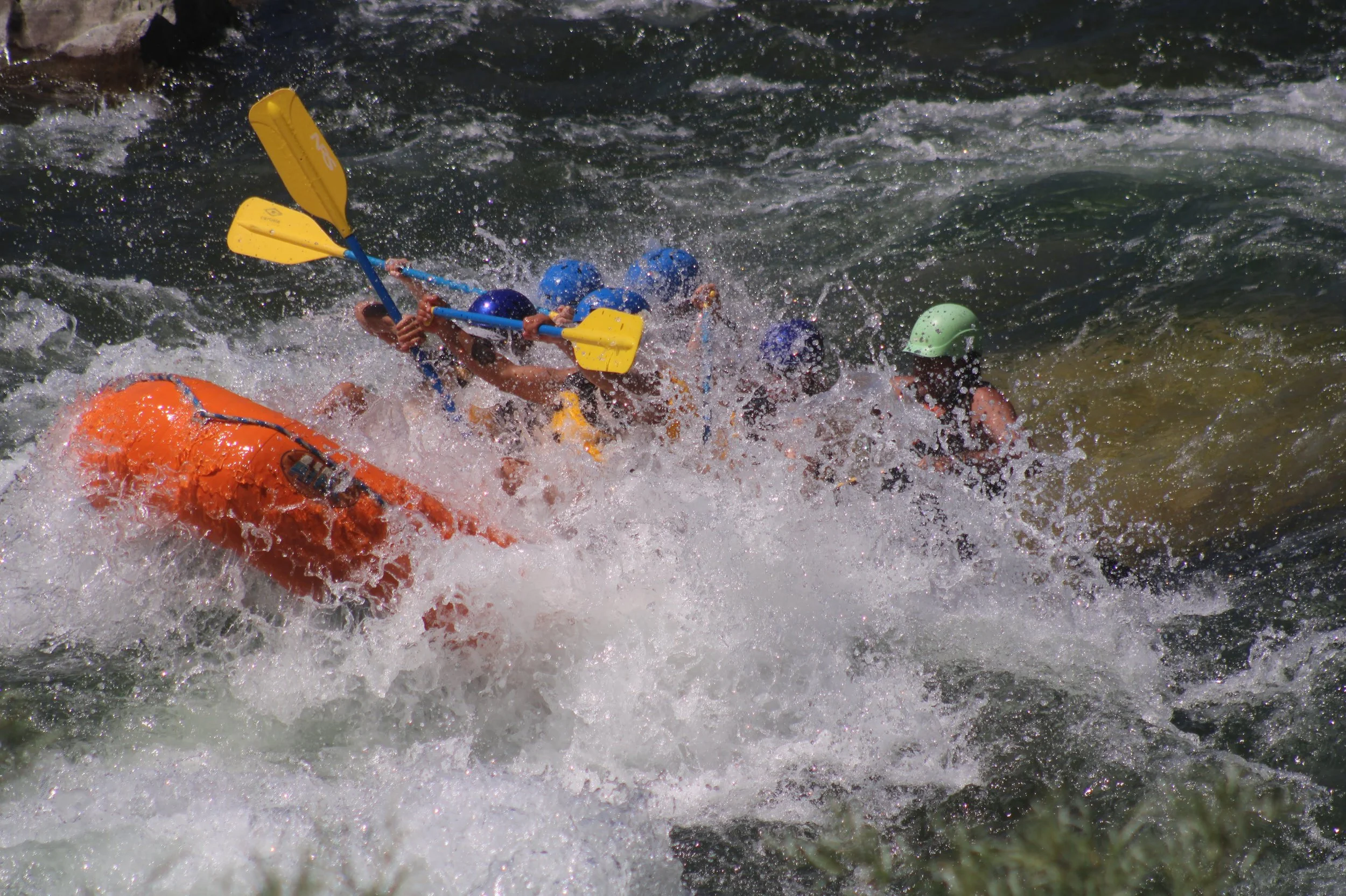Two kayakers in a rough river splash water as they pole through rapids, wearing life jackets and helmets.
