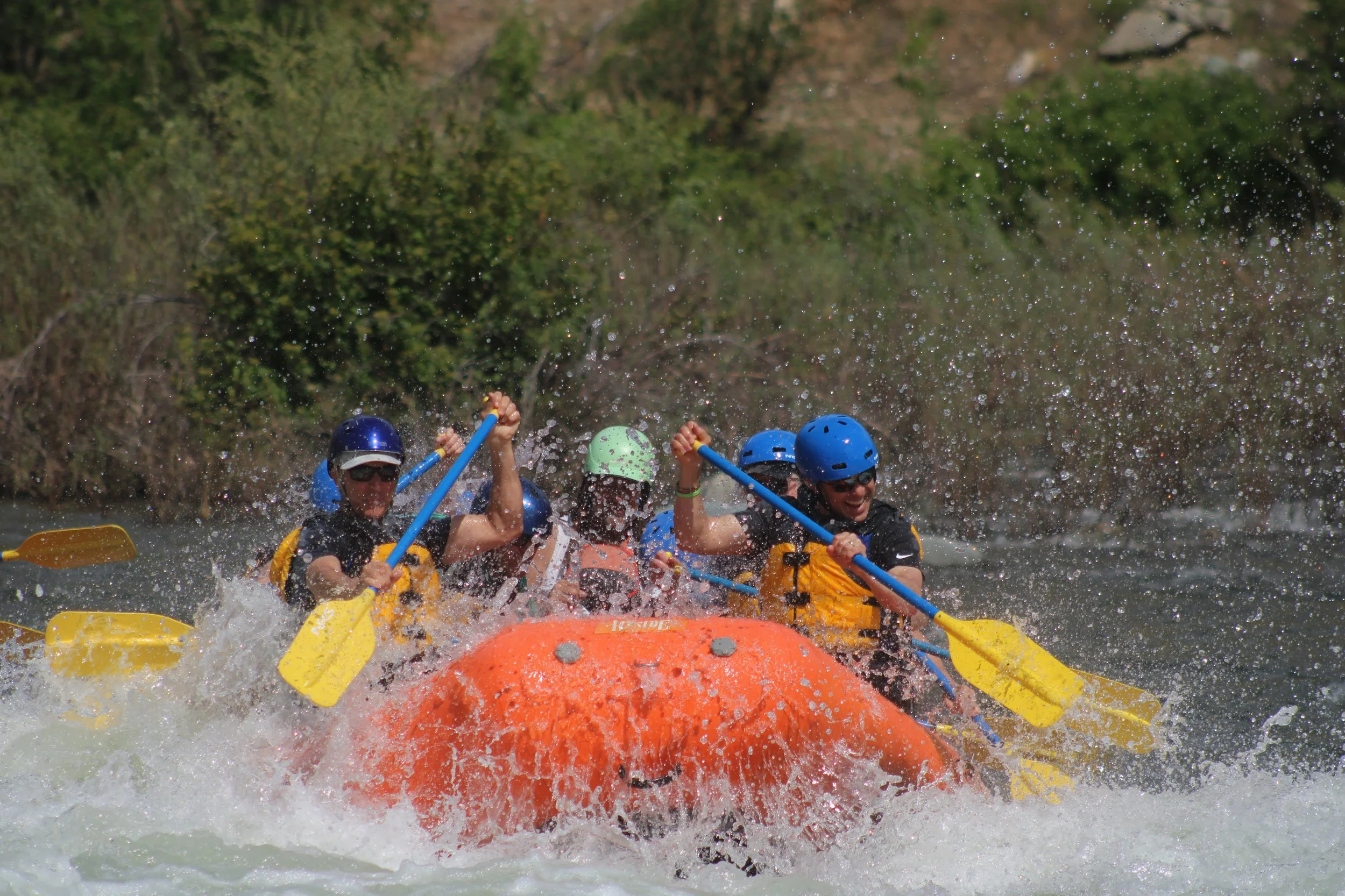 A group of six people, wearing helmets and life jackets, rafting on a river in a bright orange inflatable raft.