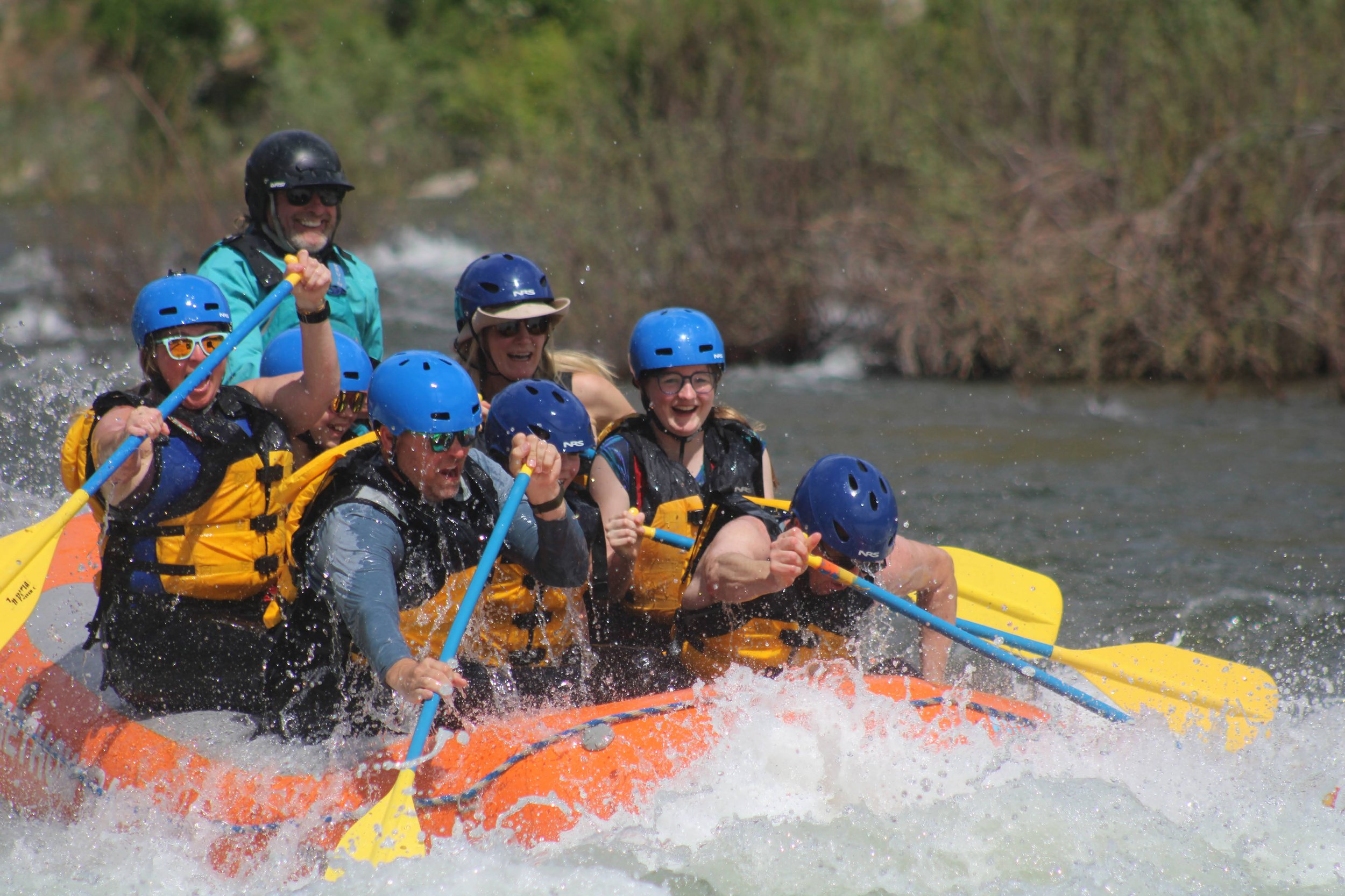 People in helmets rafting on a river with a mountainous landscape and cloudy sky in the background.
