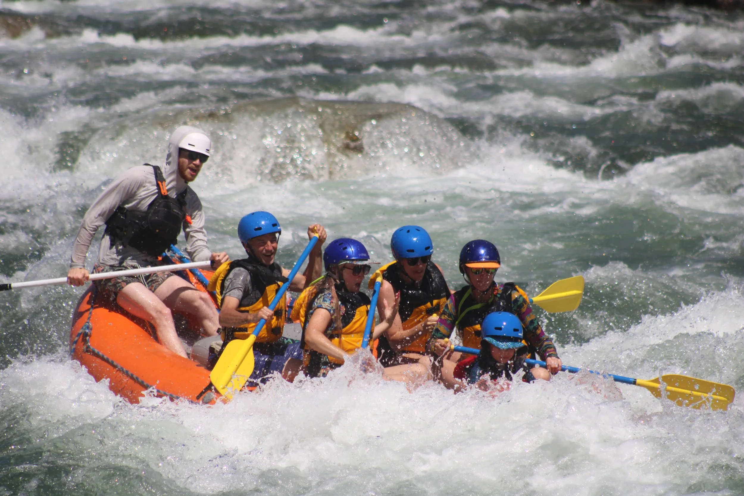 Group of six people wearing life jackets and helmets white and blue, enjoying white-water rafting on a river with rocks and trees in the background.