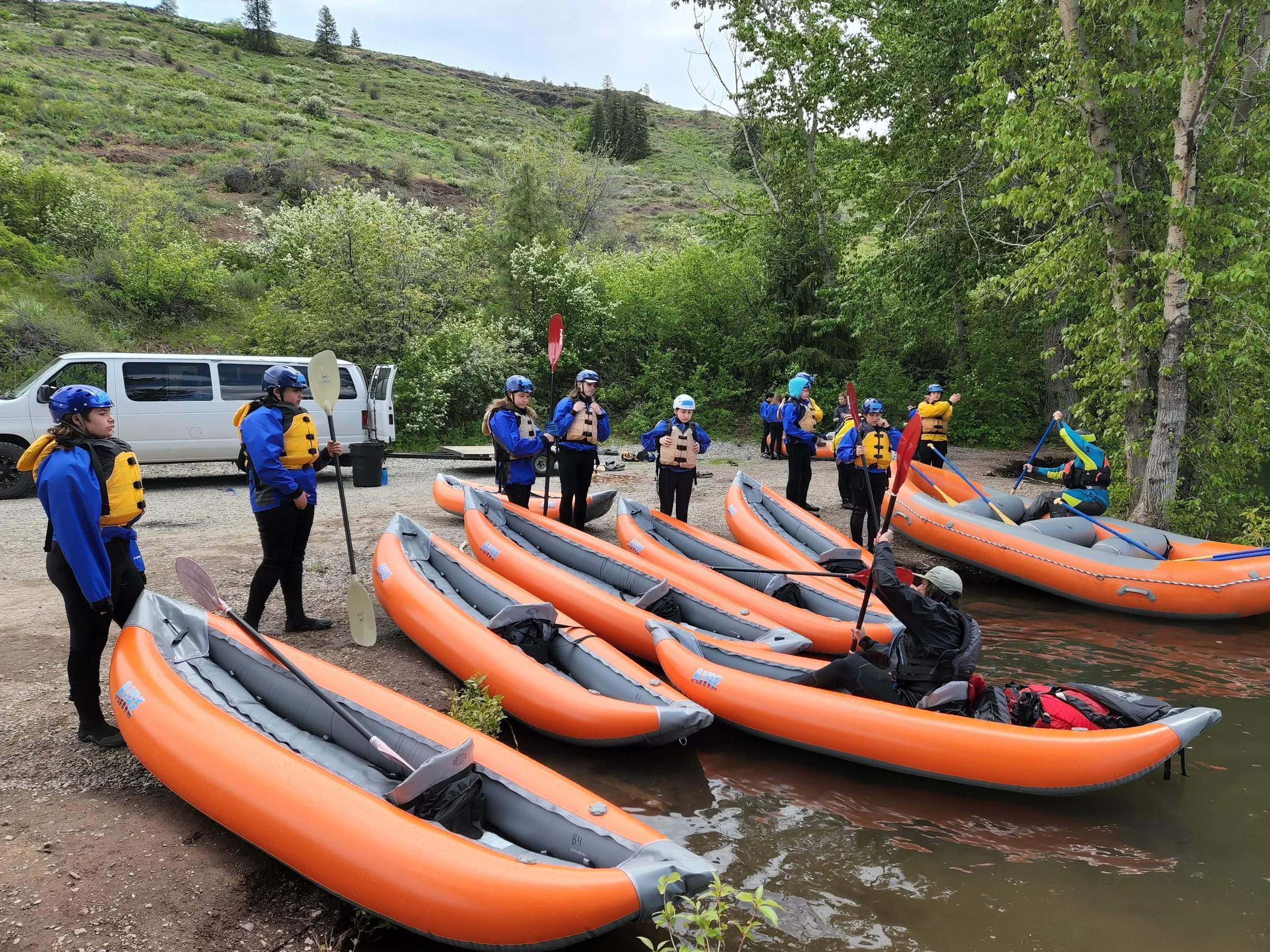 Group of people preparing for a kayaking trip beside a river, with orange inflatable kayaks, in a lush green outdoor setting.