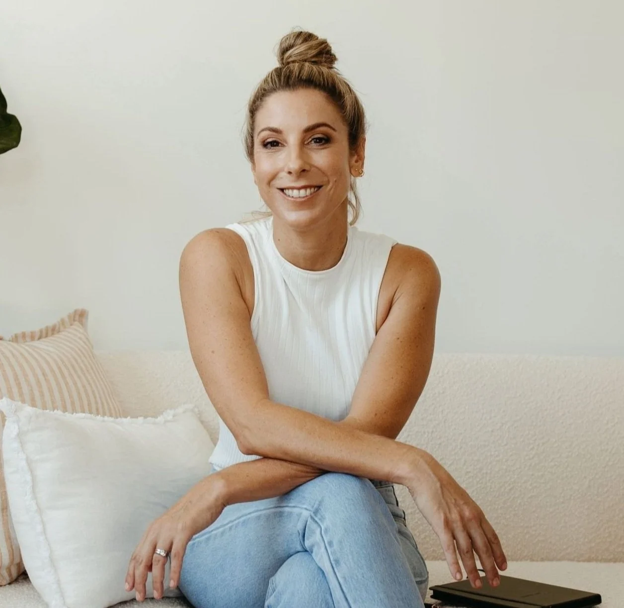 Financial marketing specialist Lisa Greensill with olive skin and dark blonde hair styled in a top bun, wearing a white sleeveless top and light blue jeans, sitting on a beige couch and smiling at the camera.