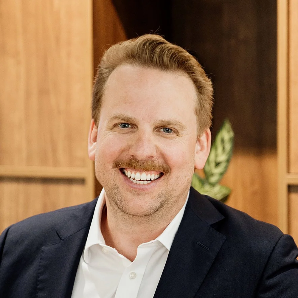 Steve Hastie from Advice Street smiling with dark ginger hair and a moustache, wearing a white shirt and black blazer, in an office setting with wooden shelves and a green plant in the background.