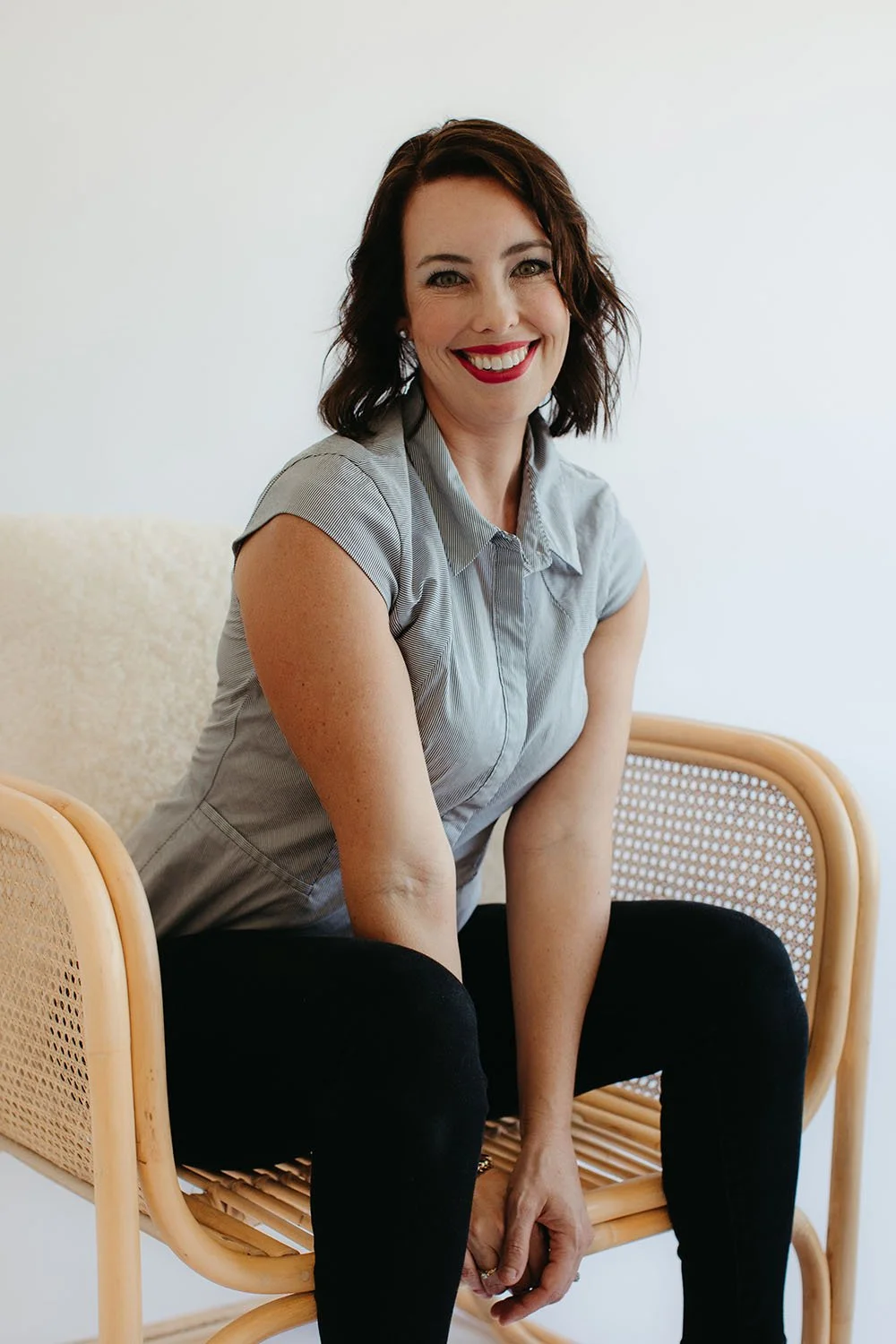 Financial brand graphic Designer and consultant Aynsley Campbell Smiling with shoulder-length dark hair and red lipstick sitting on a rattan chair, wearing a short-sleeved gray shirt and black pants, against a plain white background.