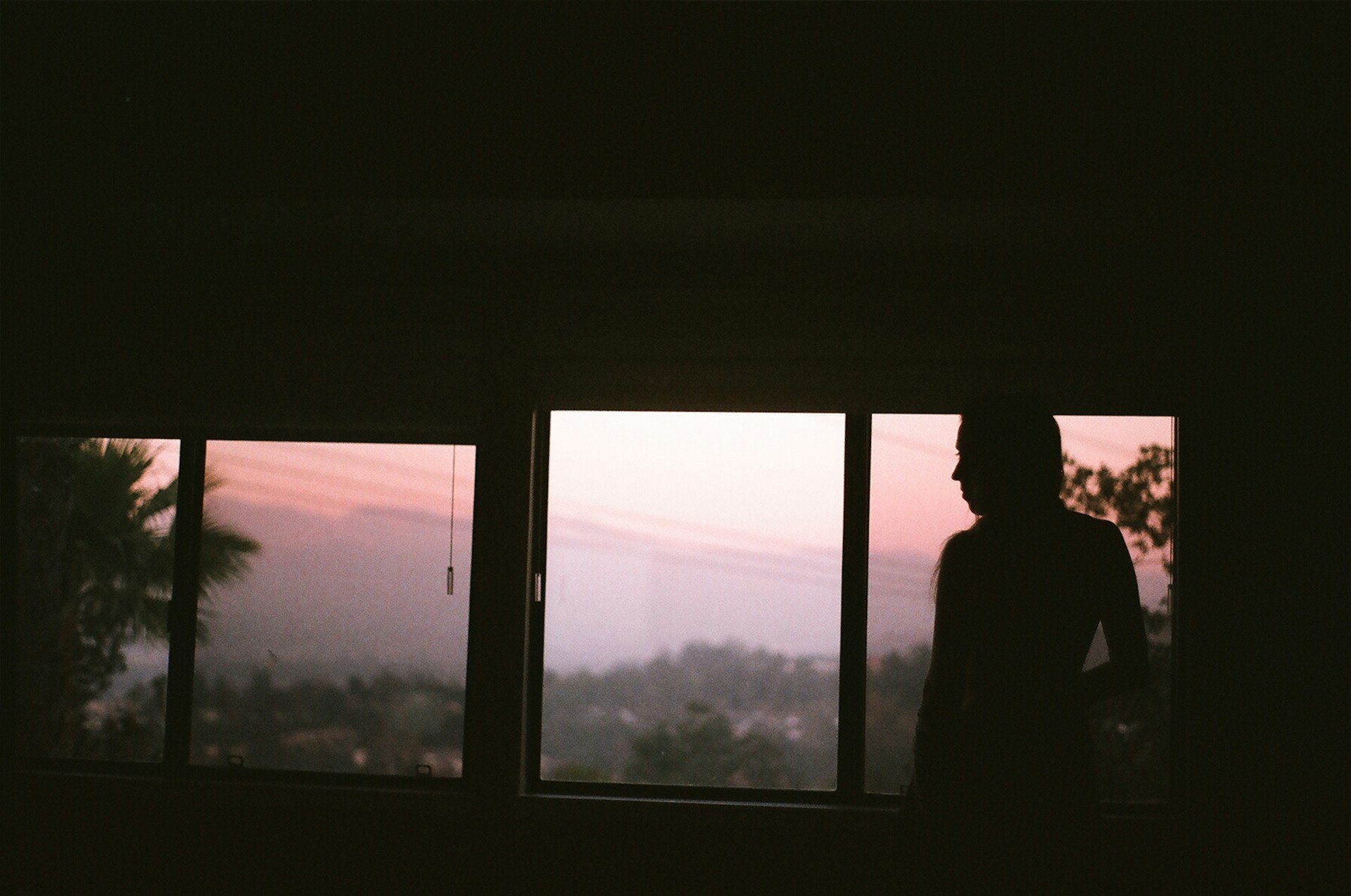 Person standing alone at a window looking out at a hazy sunset