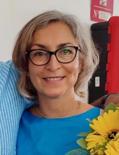 A woman with shoulder-length gray hair, wearing glasses and a blue top, smiling at the camera, with part of another person and flowers visible in the background.