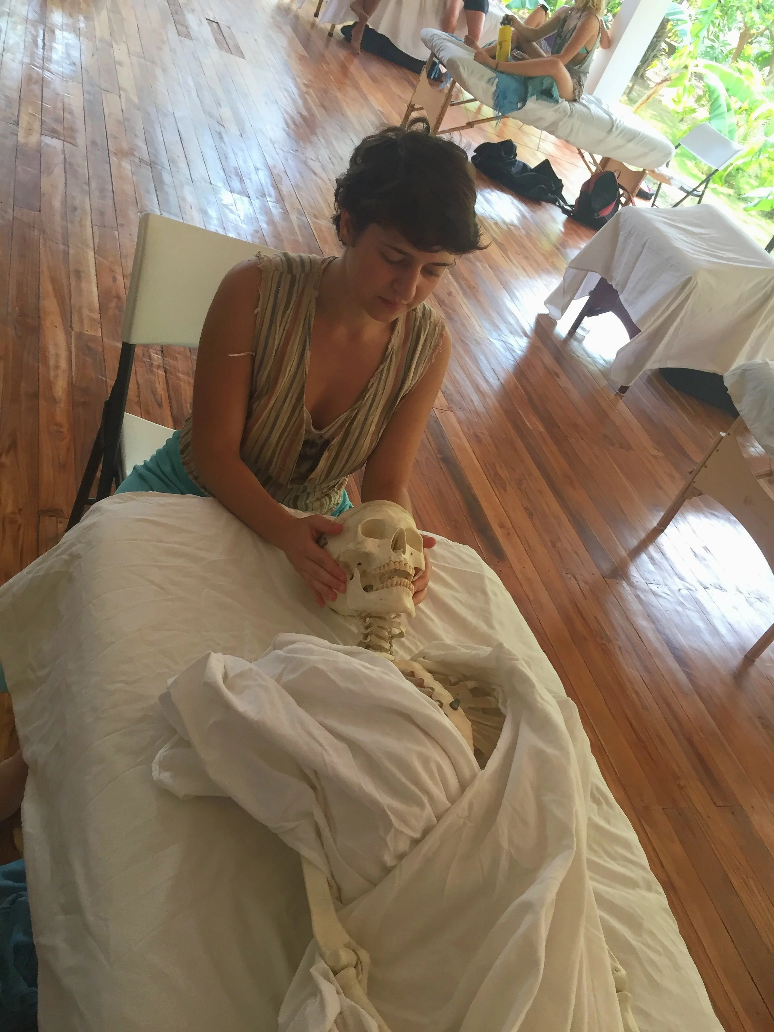 A woman with short dark hair holding a human skull and a human skull with a spine in front of her, sitting on a bed covered with white sheets in a room with wooden floors and windows with lush green foliage outside.