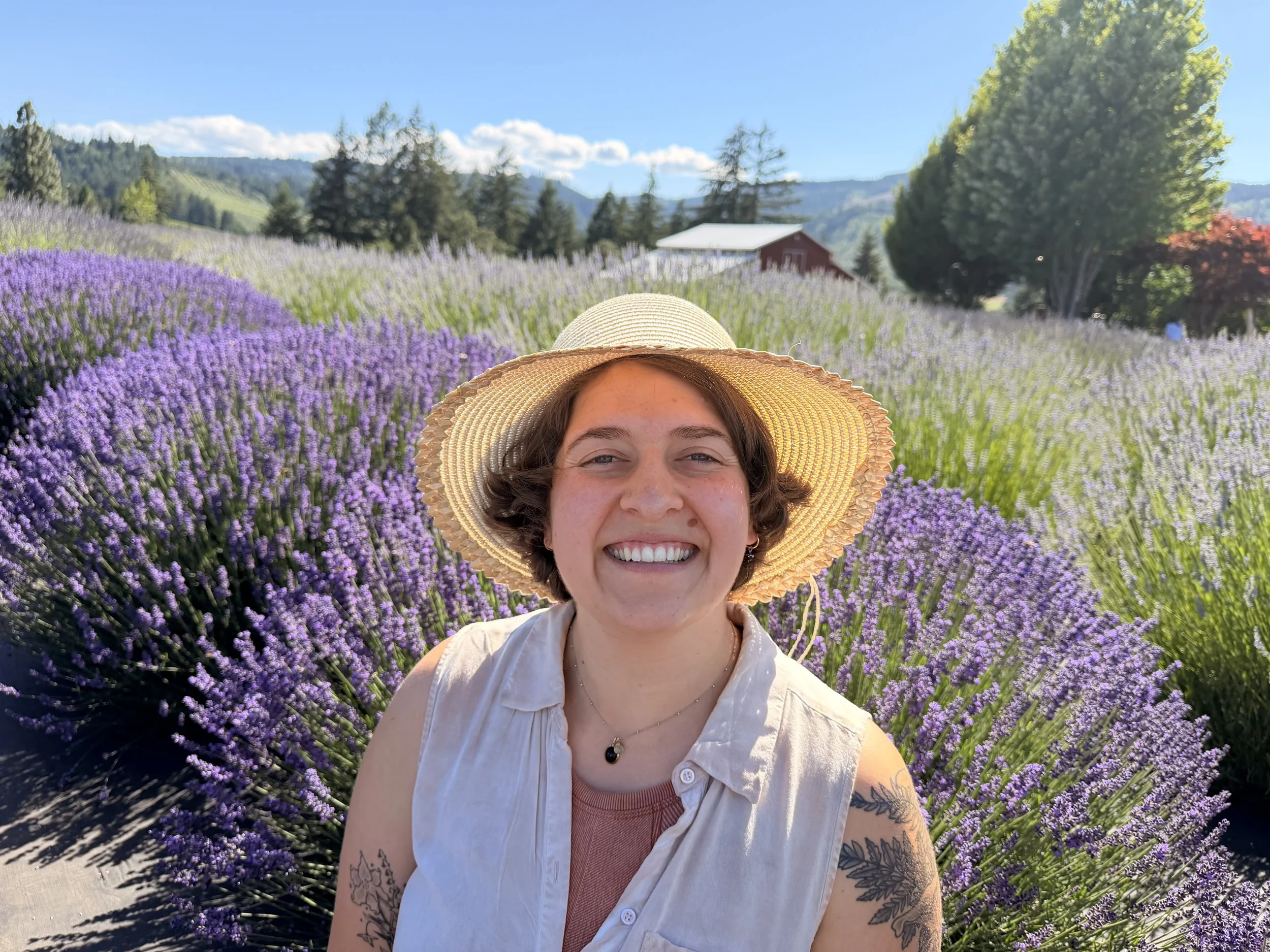 Smiling woman wearing a sunhat and sleeveless top standing in a field of purple lavender flowers during daytime.