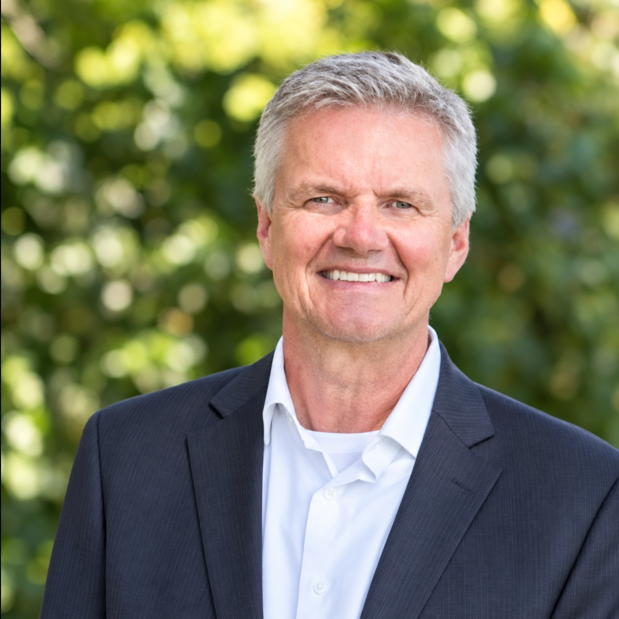 A smiling middle-aged man with gray hair, wearing a dark suit jacket and white shirt, stands outdoors with green trees in the background.