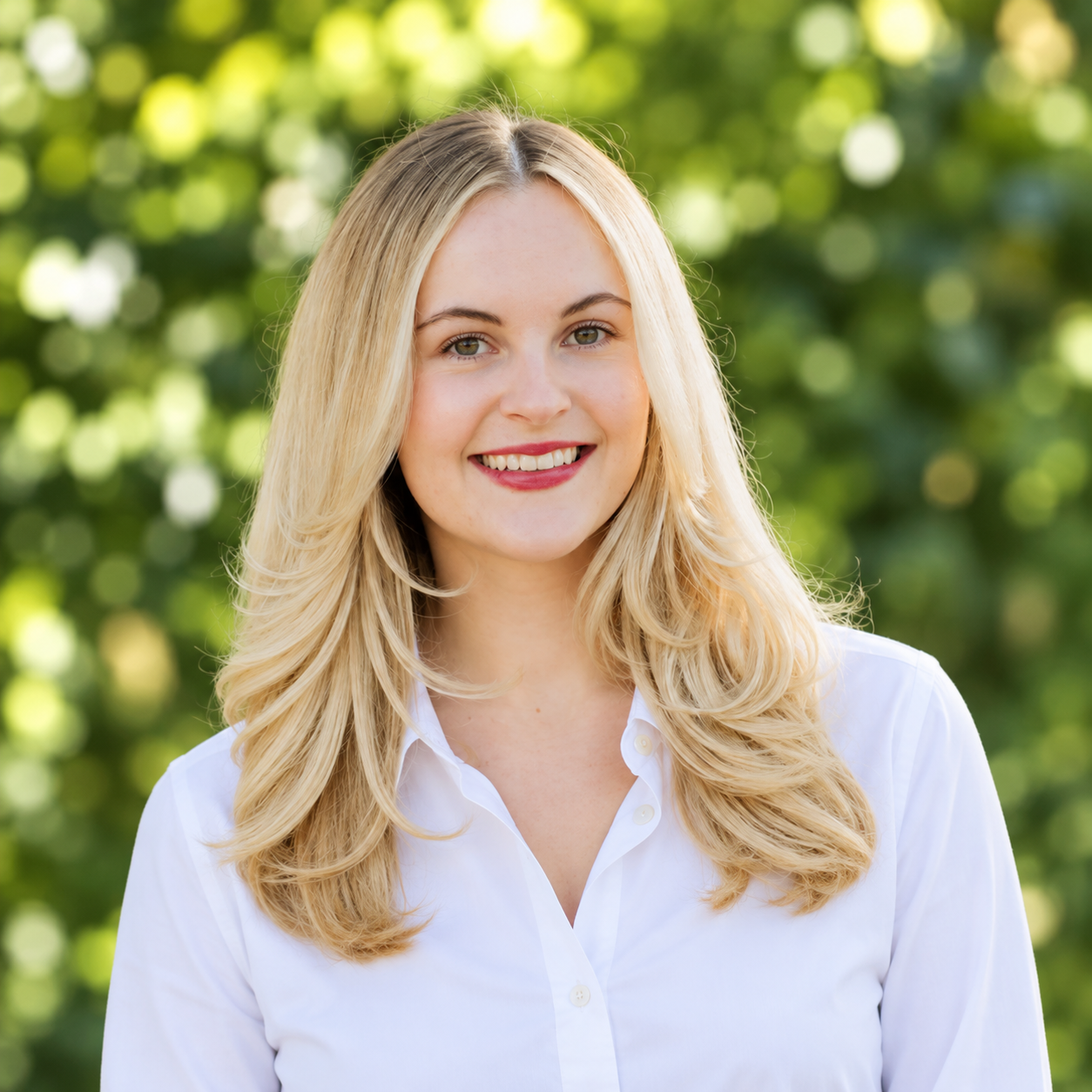 A young woman with long blonde hair, wearing a black graduation gown and white top, smiling outdoors near a stone wall with greenery in the background.