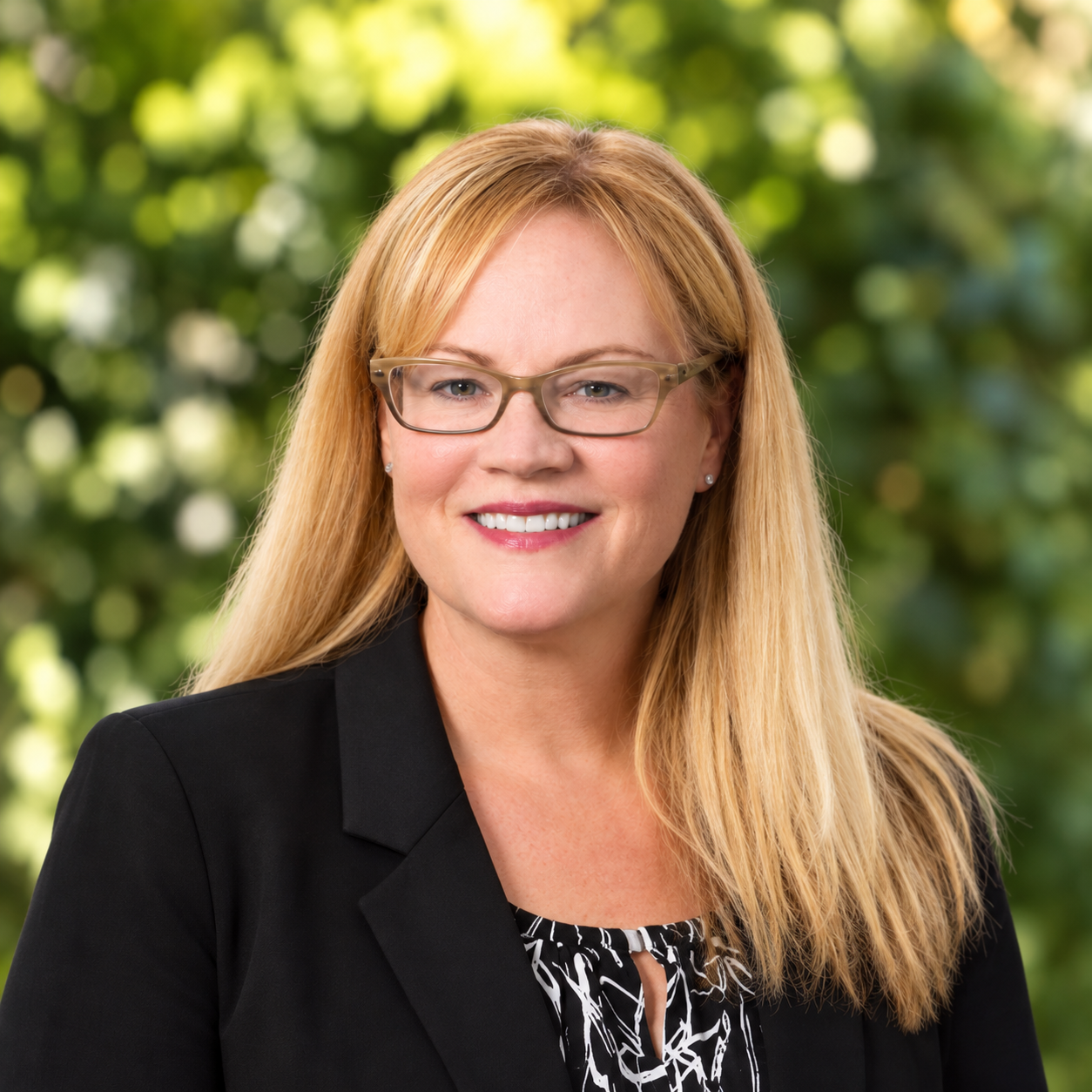 Portrait of a smiling woman with blonde hair, glasses, wearing a black blazer and patterned blouse against a gray background.