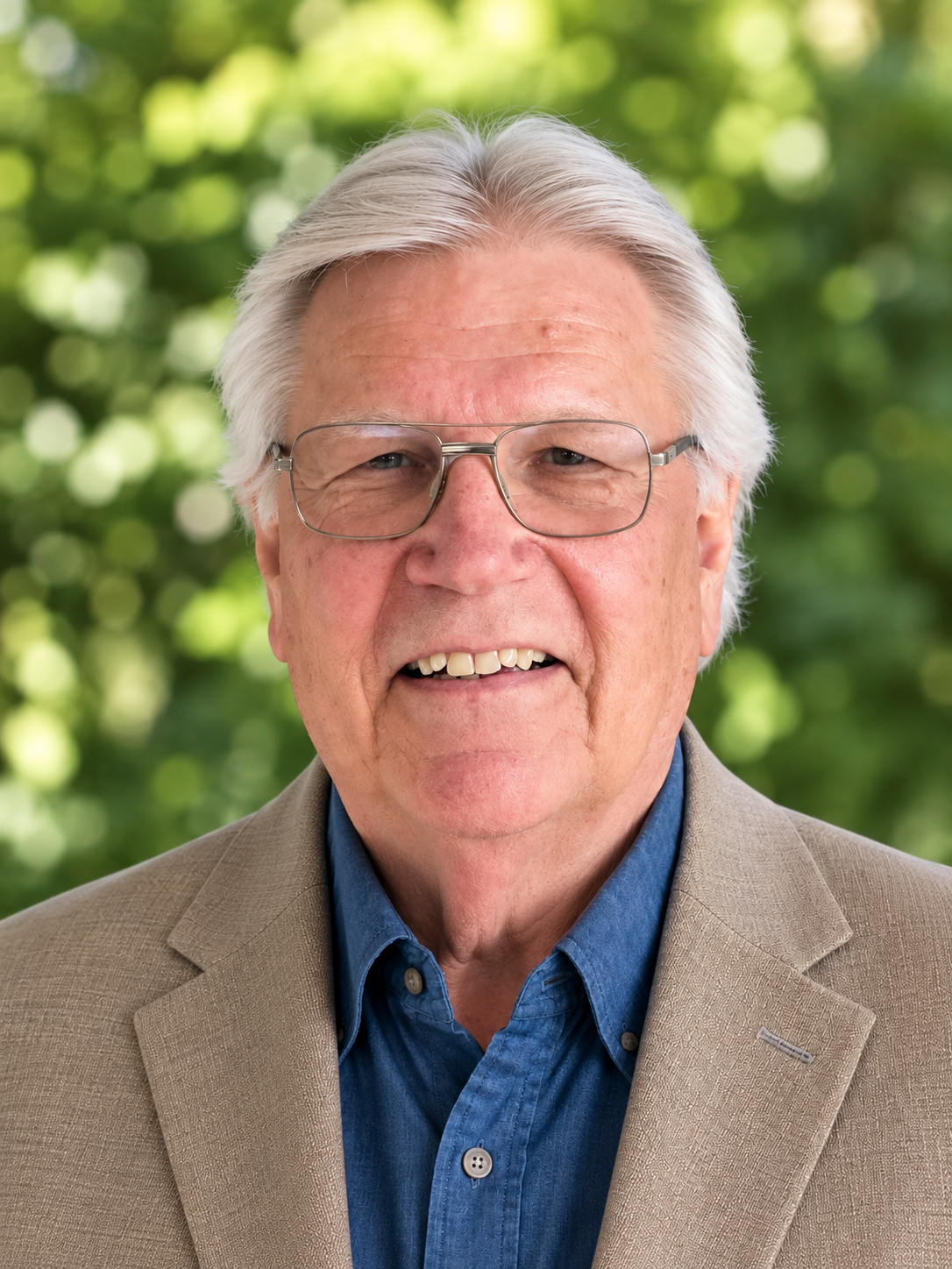A close-up photo of an elderly man with white hair, glasses, and a smile, wearing a brown blazer and a dark blue shirt, standing against a plain light-colored background.