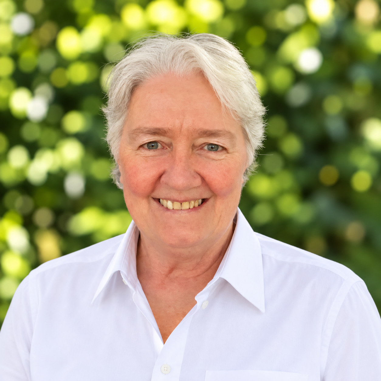 A smiling older woman with short gray hair, wearing a black, white, and gray plaid shirt and a gold necklace, standing against a dark background.