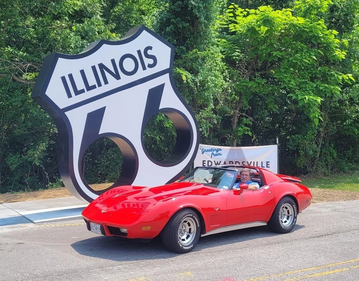 A red vintage Chevrolet Corvette convertible car parked on the road with a man in the driver's seat, near a large Illinois route 66 sign and a banner greeting from Edwardsville in the background, with trees and greenery surrounding the area.