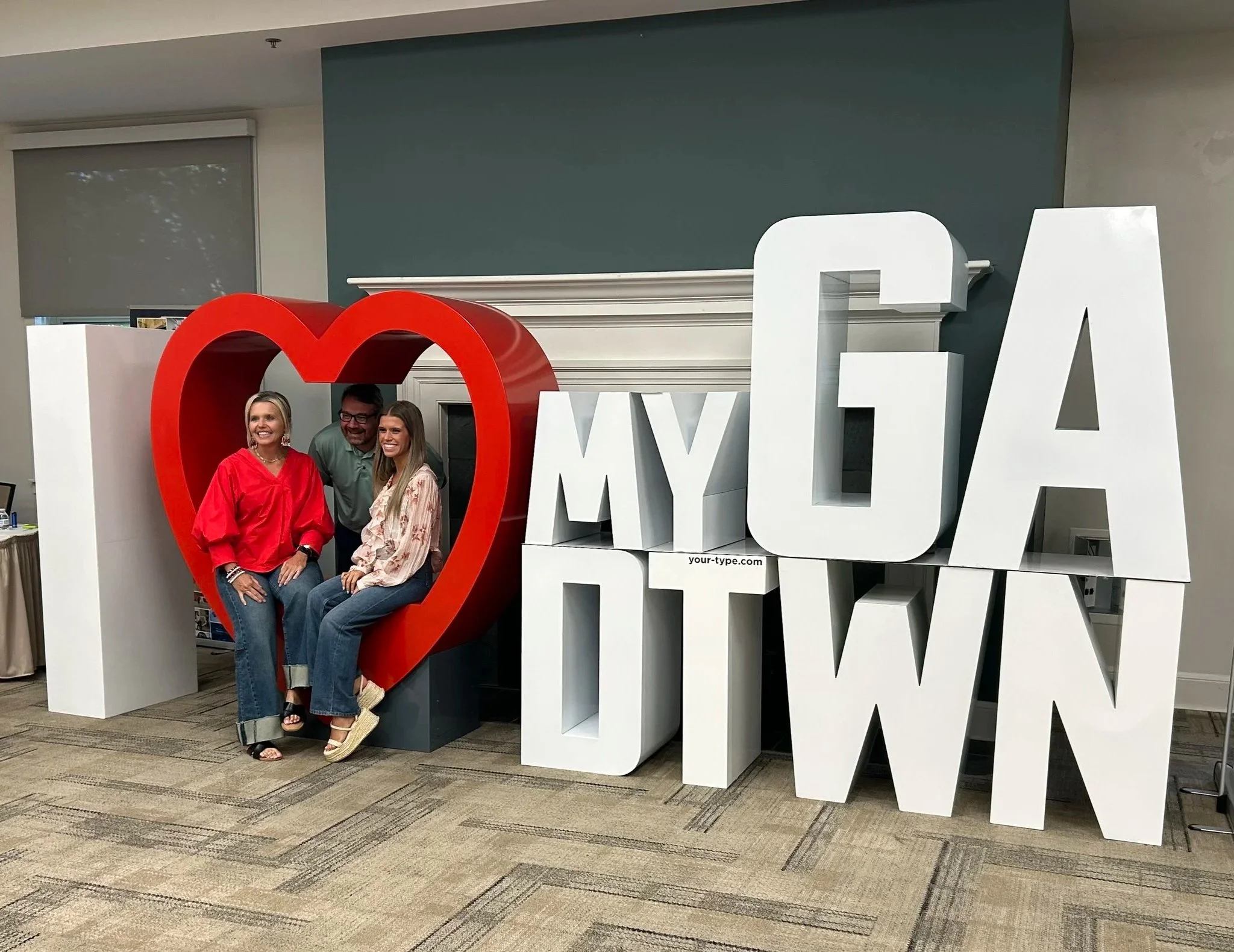Three people sitting inside a red heart-shaped display next to large white 3D letters spelling 'I ❤️ MY OA DOWN' at an indoor event.