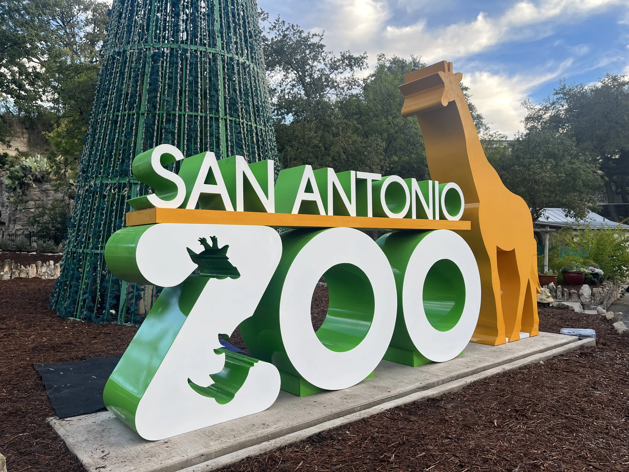 Colorful sign reading 'San Antonio 200' with a giraffe figure on the right side, outdoor setting with trees and cloudy sky.