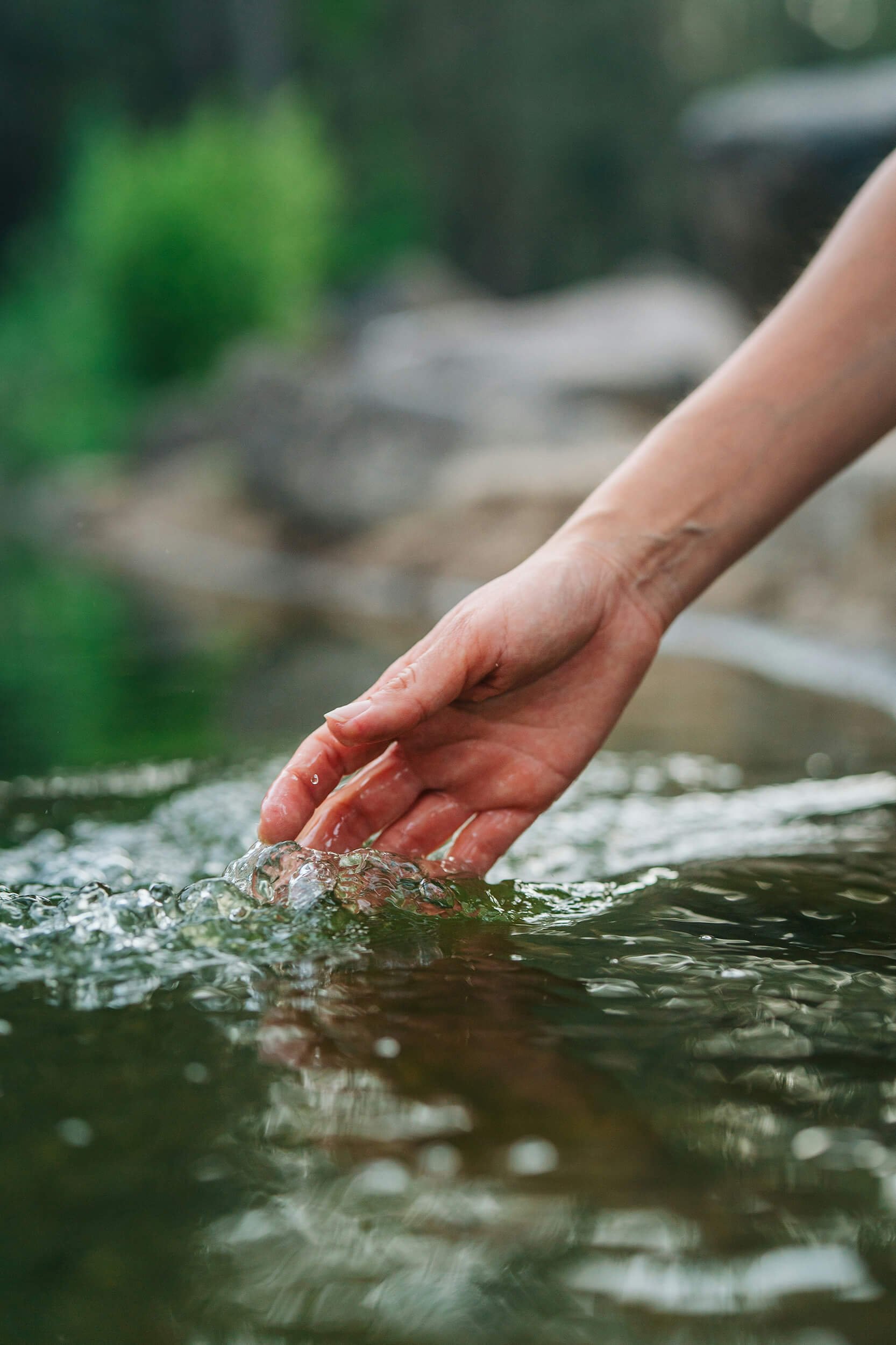 A pair of hands submerged in shallow water, gently touching the surface and creating small ripples. The background is blurred with hints of green foliage.