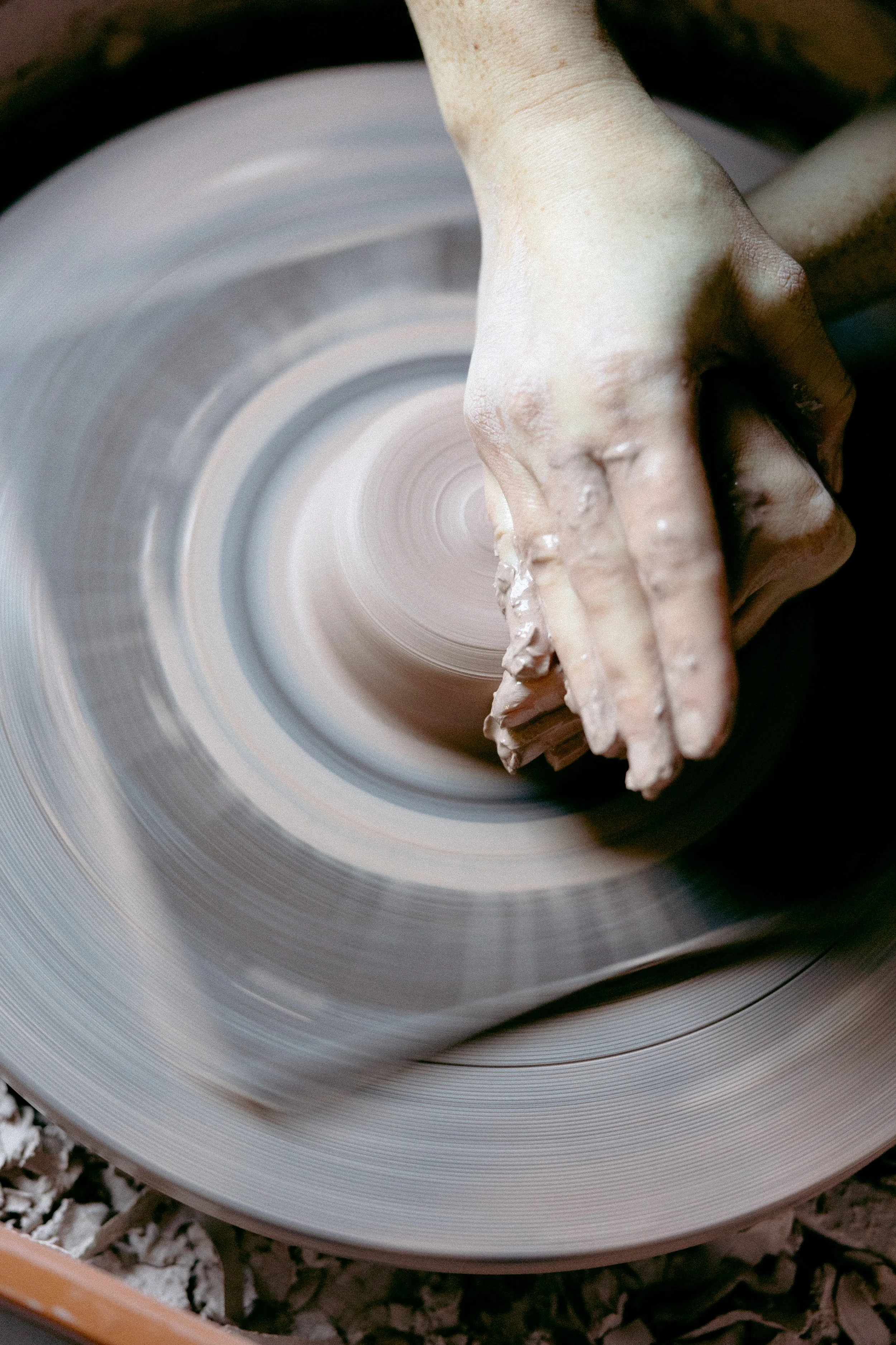 A person shaping clay on a spinning pottery wheel, with visible clay on their hands and a blurred background.