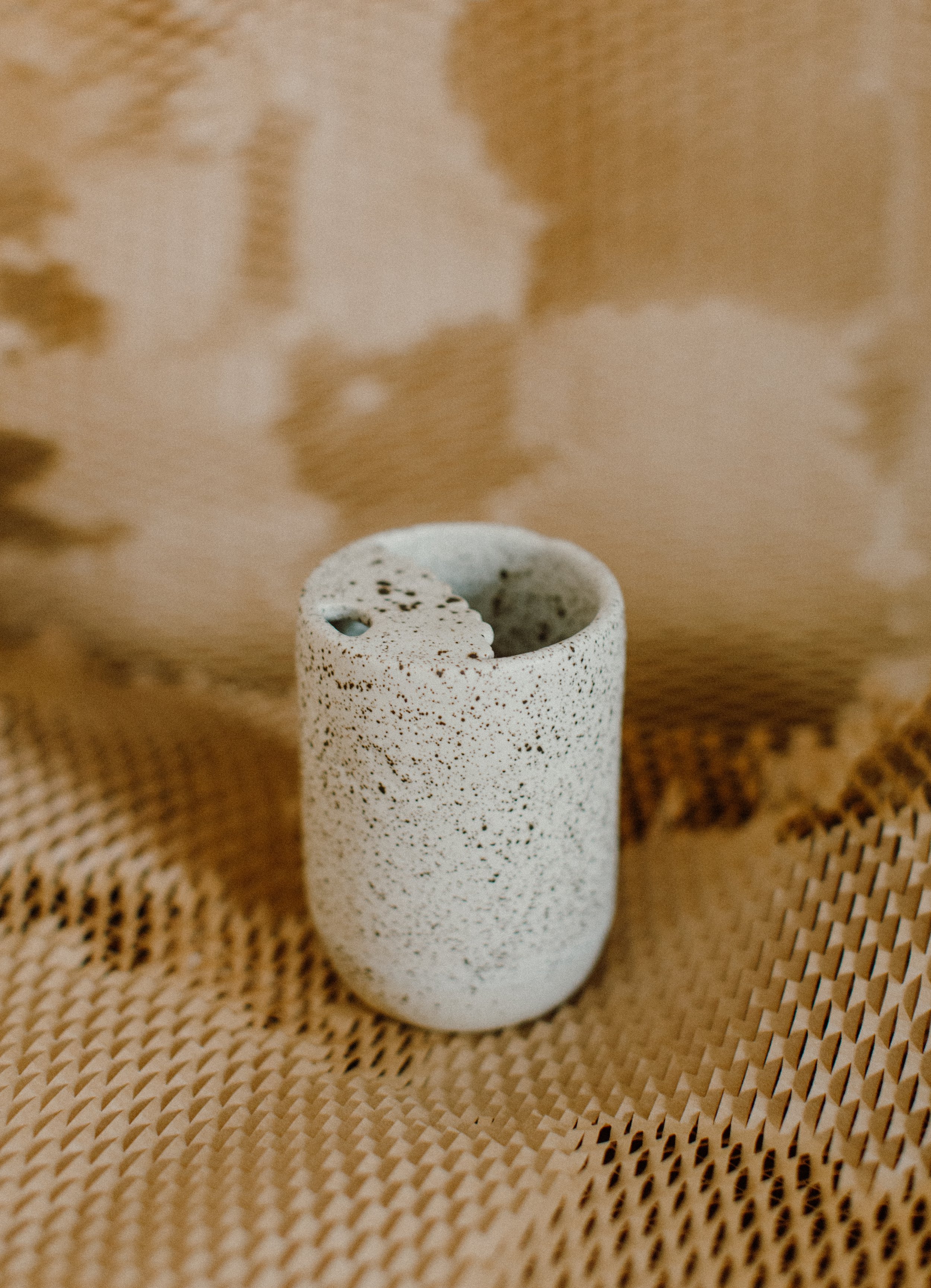 A speckled white ceramic vase with a small opening at the top, placed on a textured woven surface.