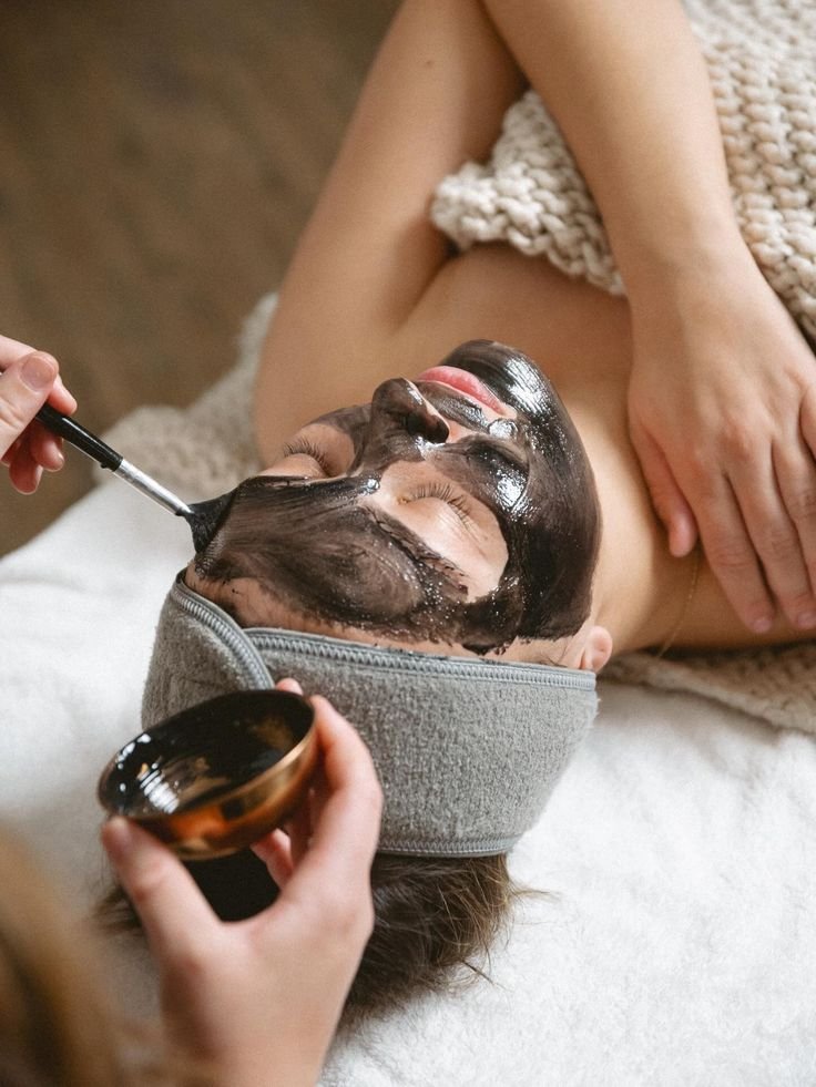 Person lying down wearing green protective goggles and a red and blue striped shirt, with a black and gray face mask or skin covering, in a medical or spa setting.