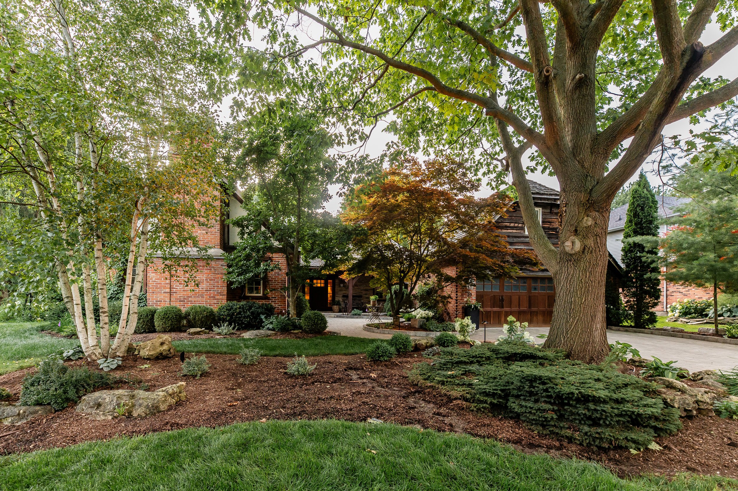 Front yard with lush landscaping, brick house, large trees, and driveway.