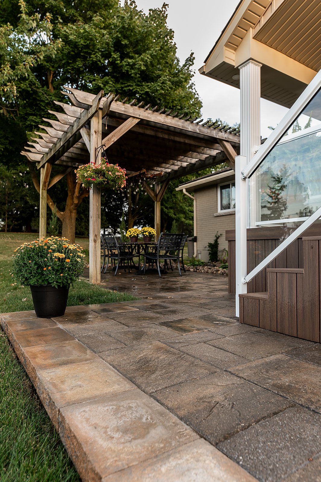 Outdoor patio with wooden pergola, hanging flowers, and outdoor dining set on stone paving, adjacent to house stairs.